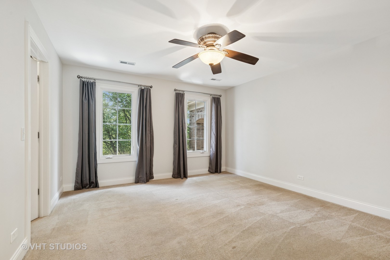 Empty carpeted bedroom with two curtained windows and a ceiling fan with light.