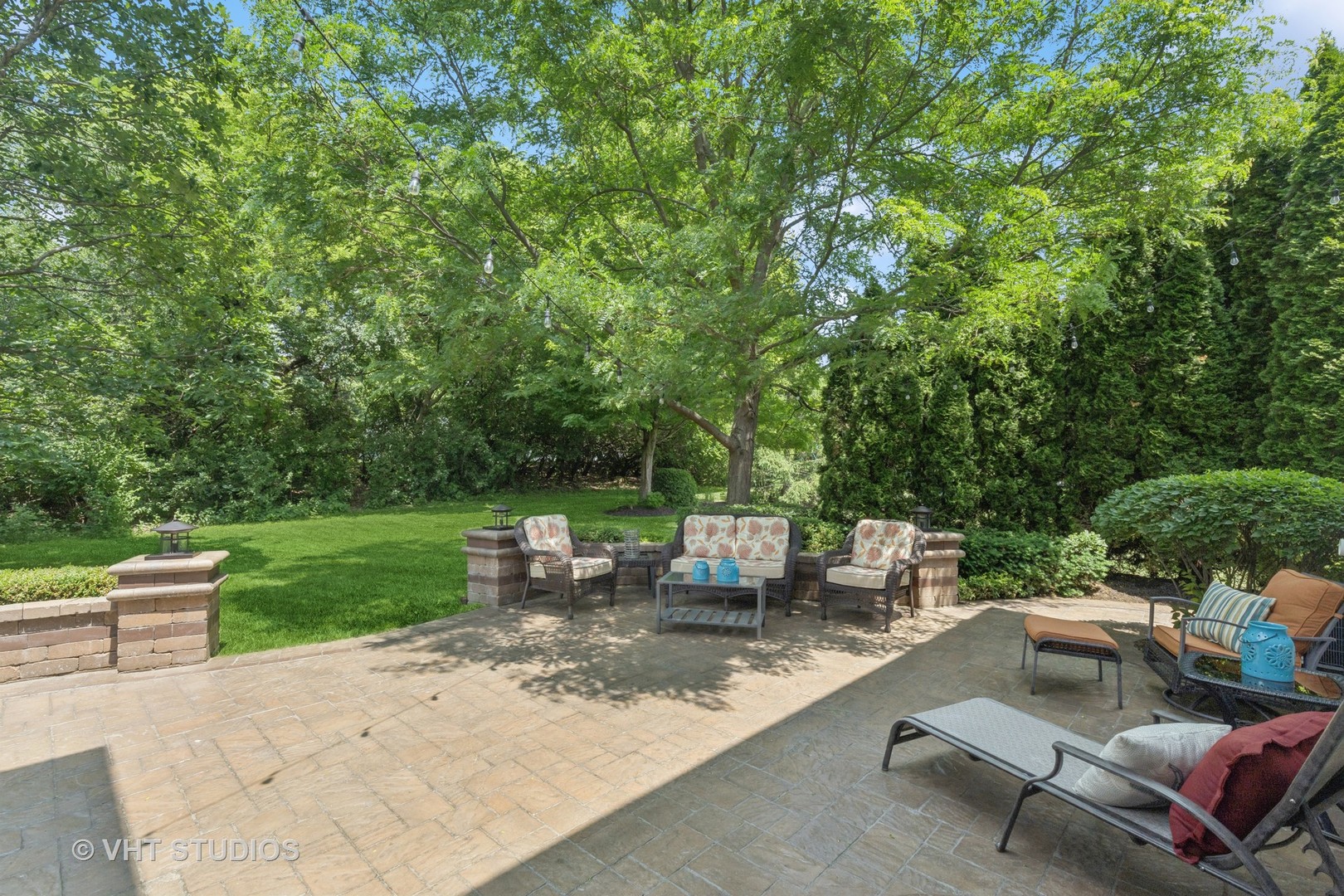 Backyard patio with wicker seating, stone pavers, mature trees, and evergreen hedges.