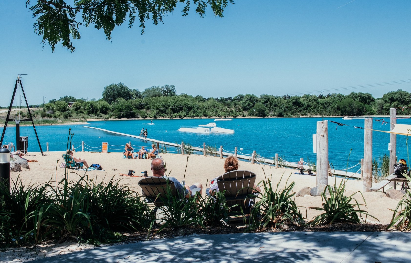 Sandy lakeside beach with people relaxing and a floating wakeboarding obstacle on the water.