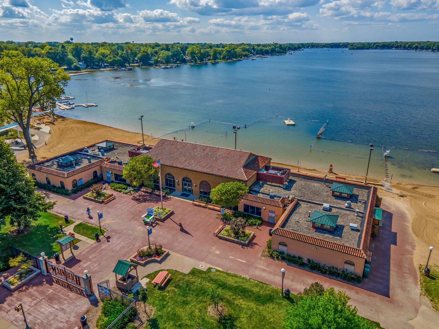 Lakeside clubhouse and sandy beach beside a buoy-marked swimming area and docks.