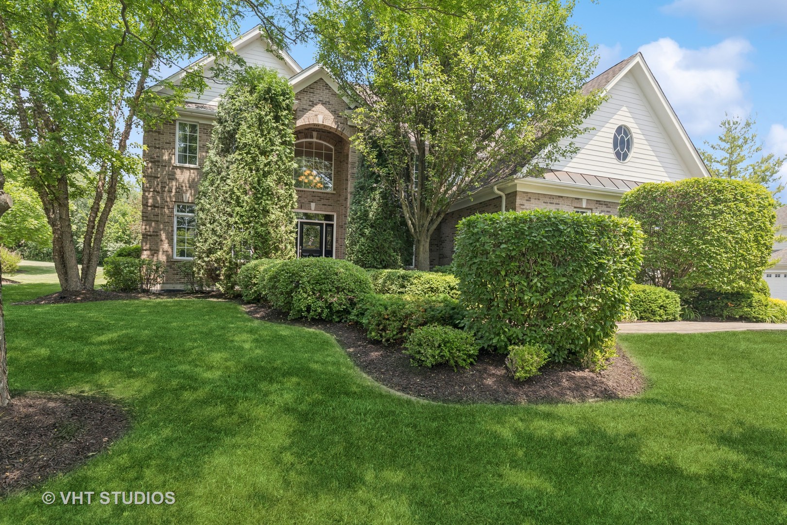 Two-story brick home with white gables, a circular window, mature trees, and manicured landscaping.