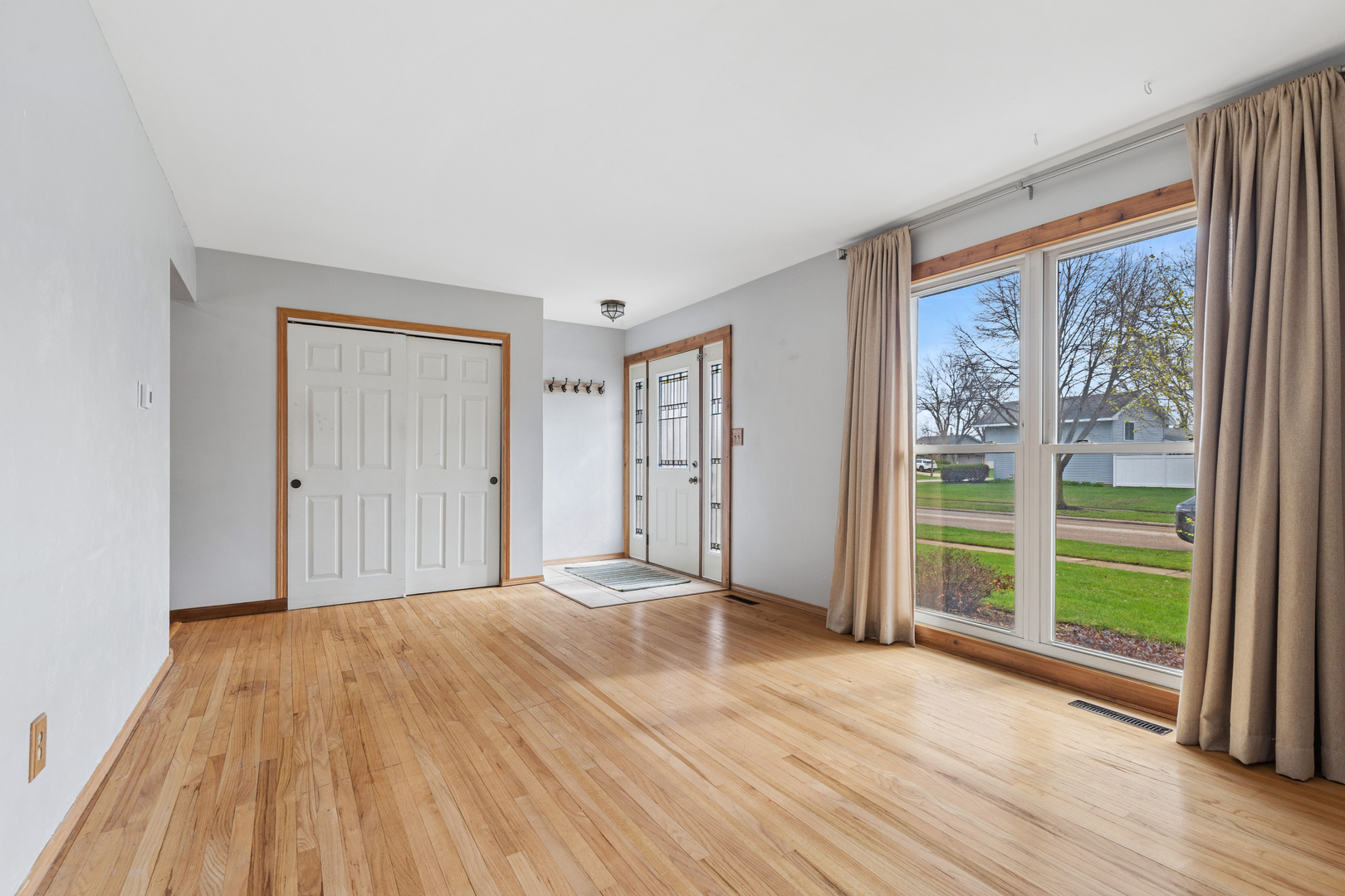 Empty living room and entry with hardwood floors, decorative glass sidelights, and a large curtained window.
