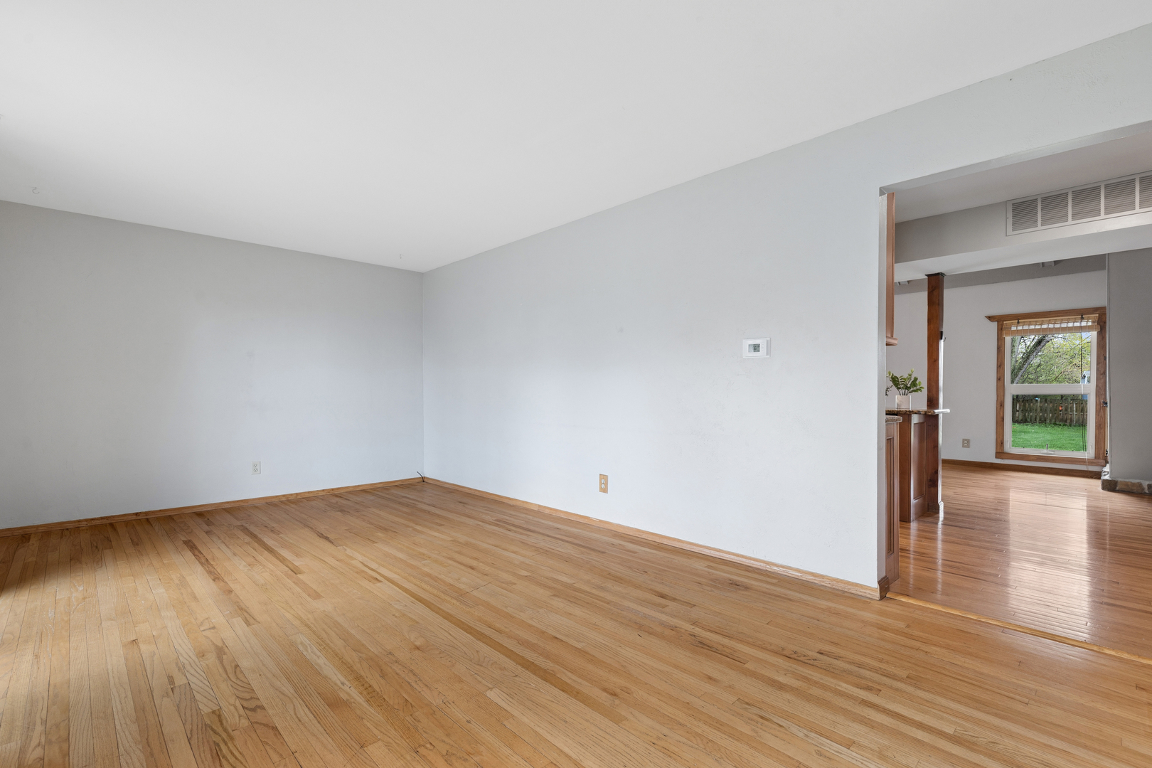 Empty living room with hardwood floors and an open doorway to a room with a wood-trimmed window.