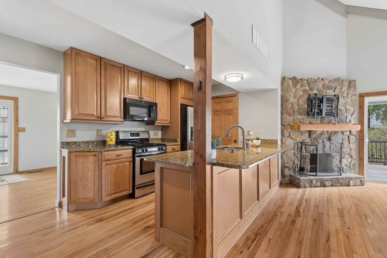 Kitchen with wood cabinets and granite island beside a stone fireplace and hardwood floors.