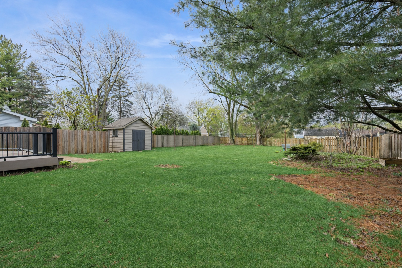 Fenced backyard with a broad lawn, detached shed, deck, and mature trees.