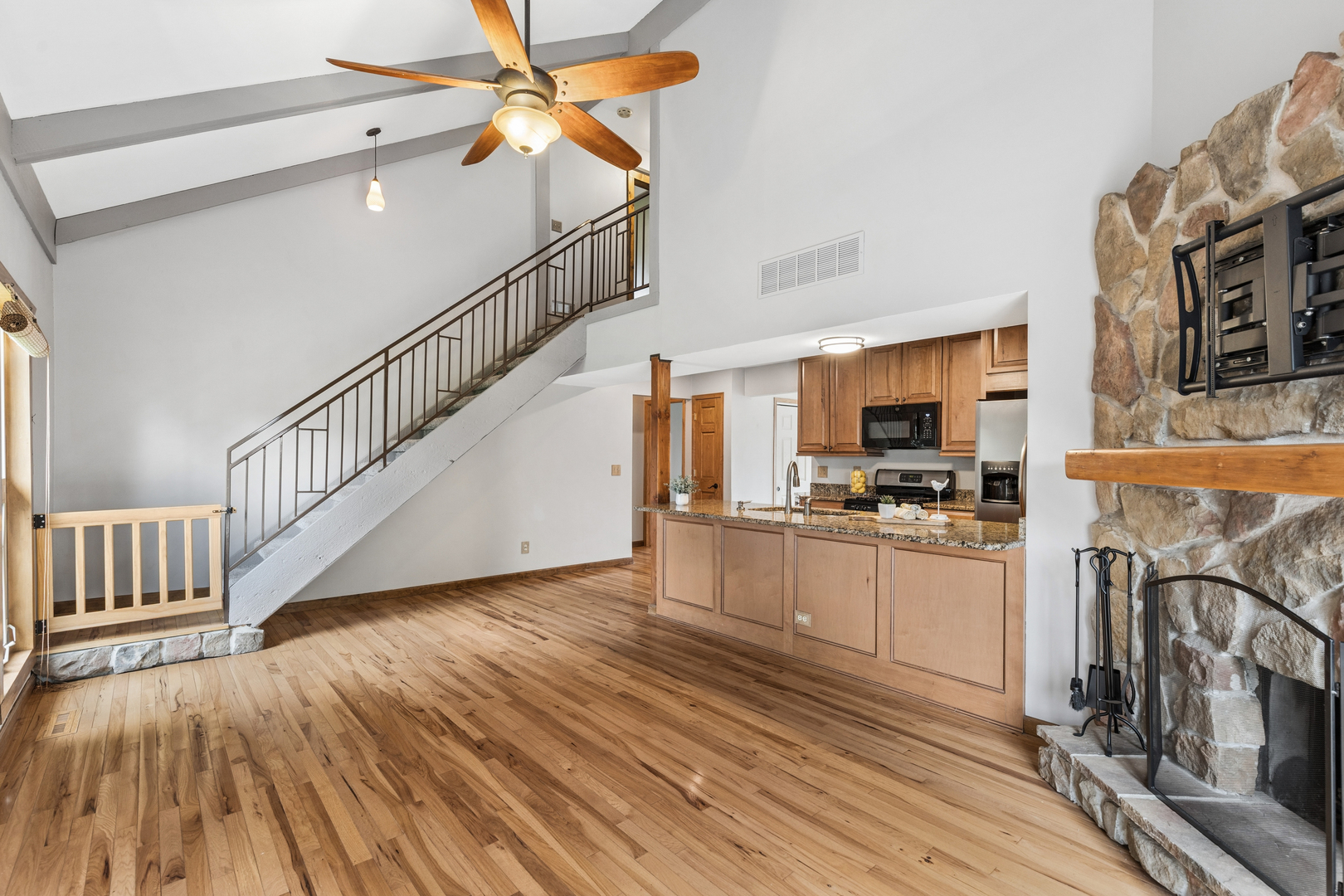 Open living room with hardwood floors, stone fireplace, staircase, and adjoining kitchen.