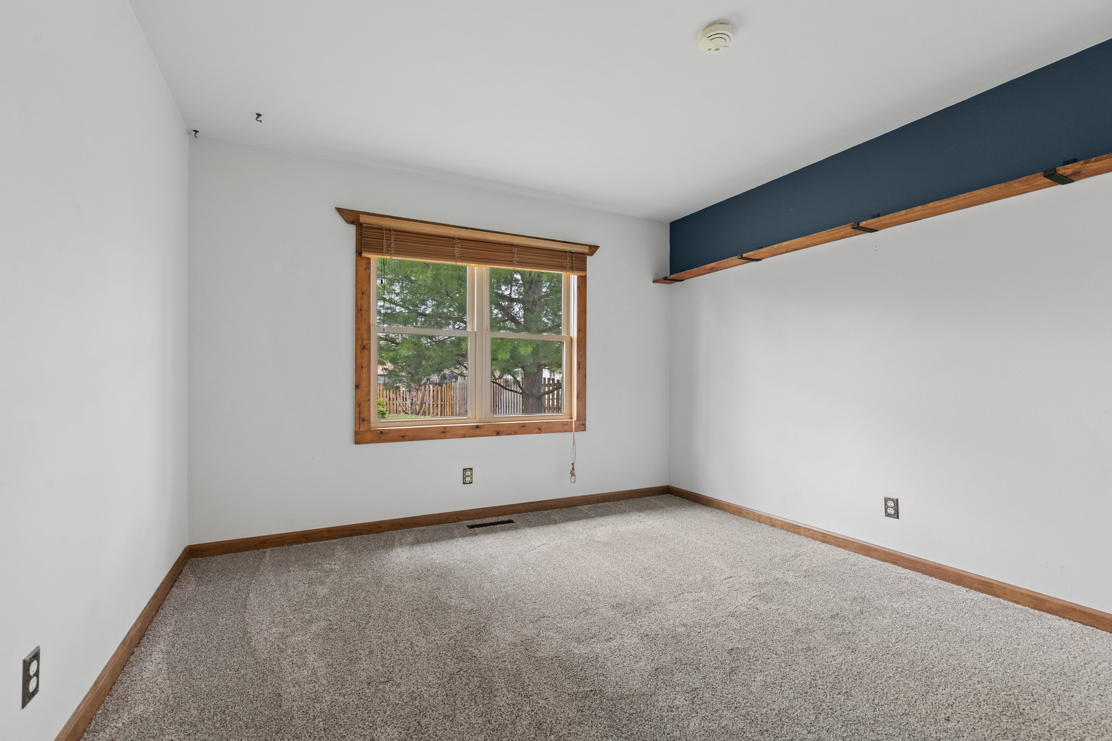 Empty carpeted room with a wood-trimmed window and long shelf along a dark blue accent wall.