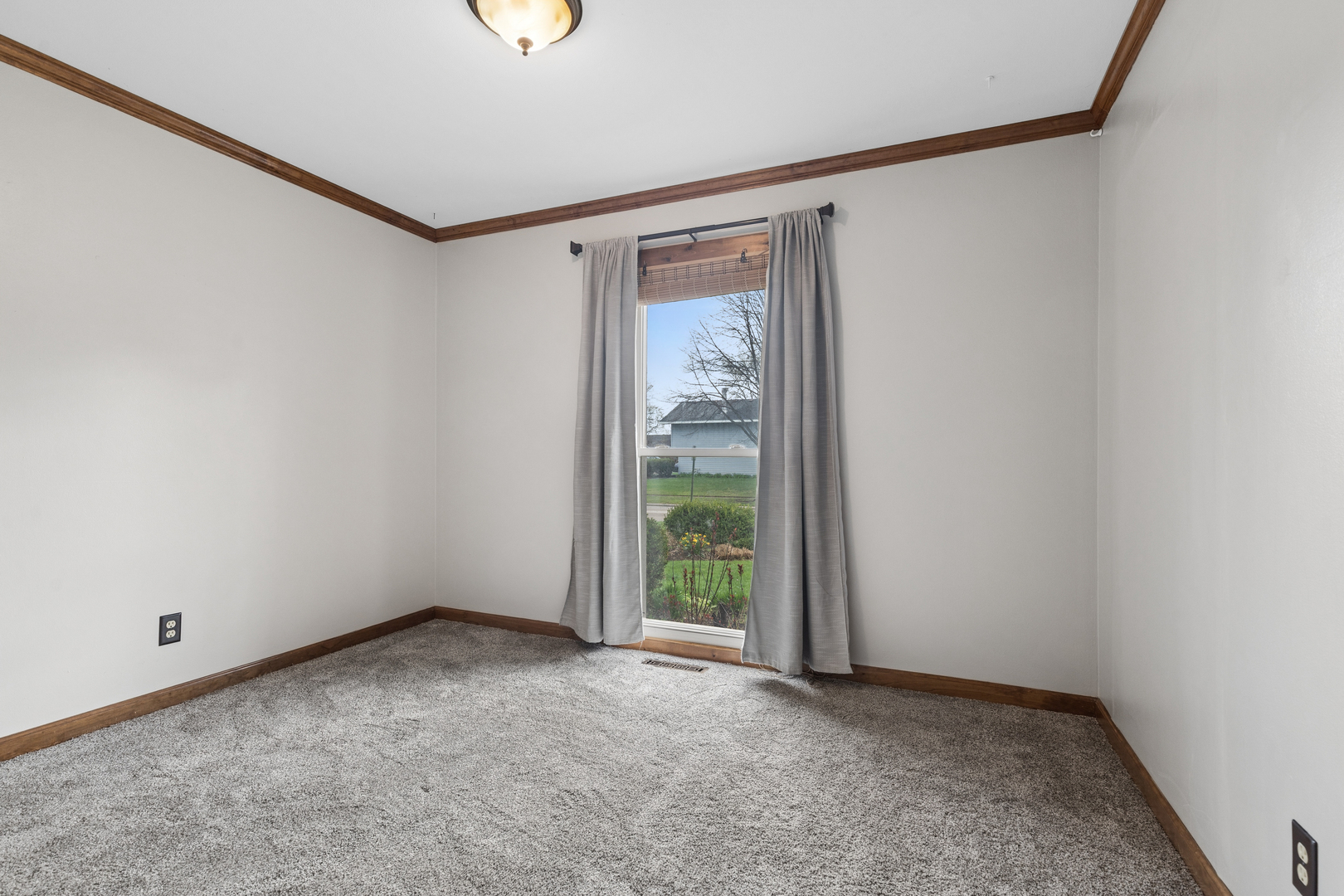 Empty carpeted bedroom with dark wood trim and a curtained window overlooking the yard.