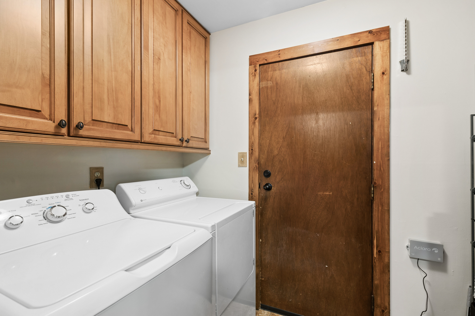 Laundry room with white washer and dryer beneath wood cabinets beside a wood-trimmed door.
