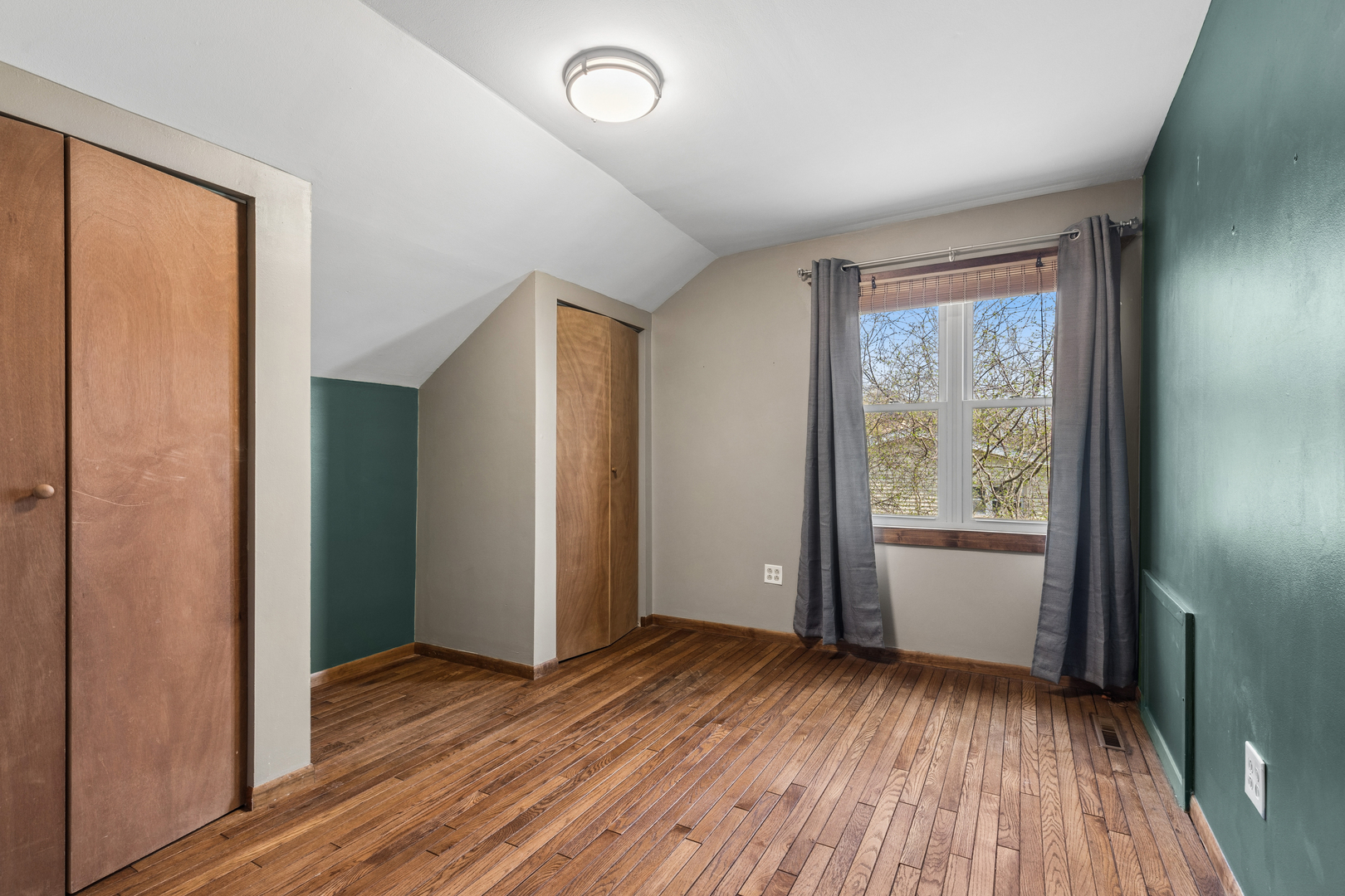 Unfurnished bedroom with hardwood floors, built-in closets, a sloped ceiling, and gray curtains framing the window.