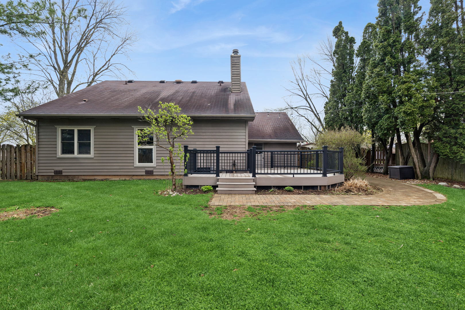 Rear of gray-sided house with raised deck, black railings, curved paver walkway, and spacious lawn.