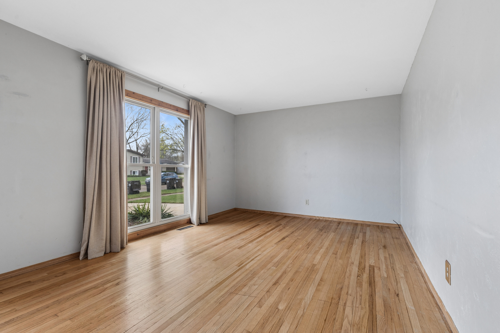 Empty room with hardwood floors and a large curtained window overlooking the street.