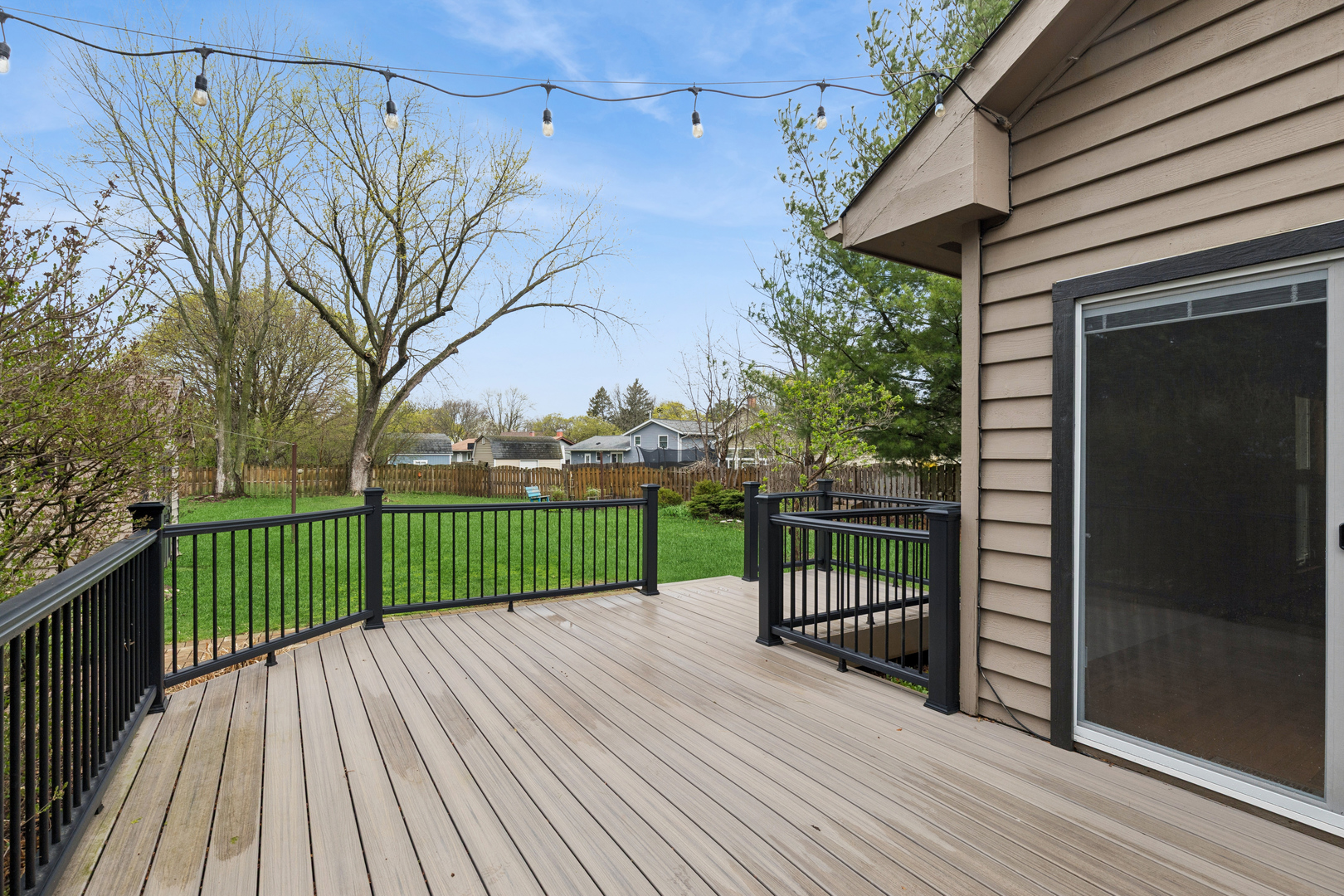 Elevated wood deck with black railings overlooking a fenced backyard, with string lights overhead.