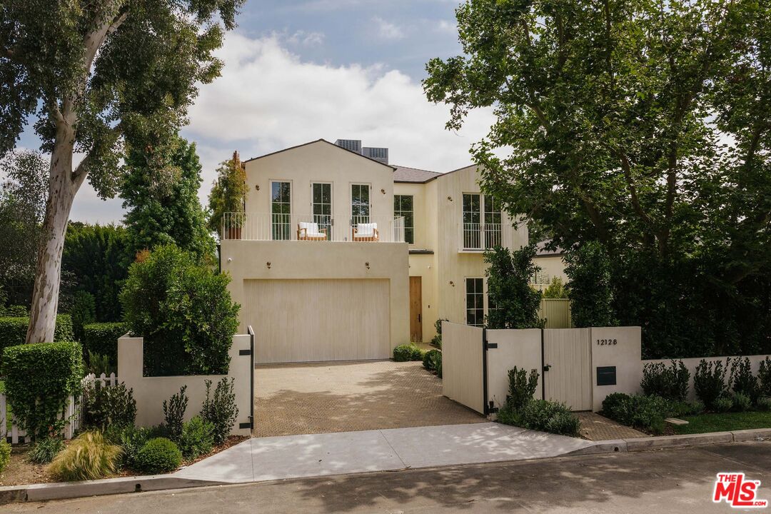 Modern cream two-story home with tall windows, a gated driveway, and mature trees.