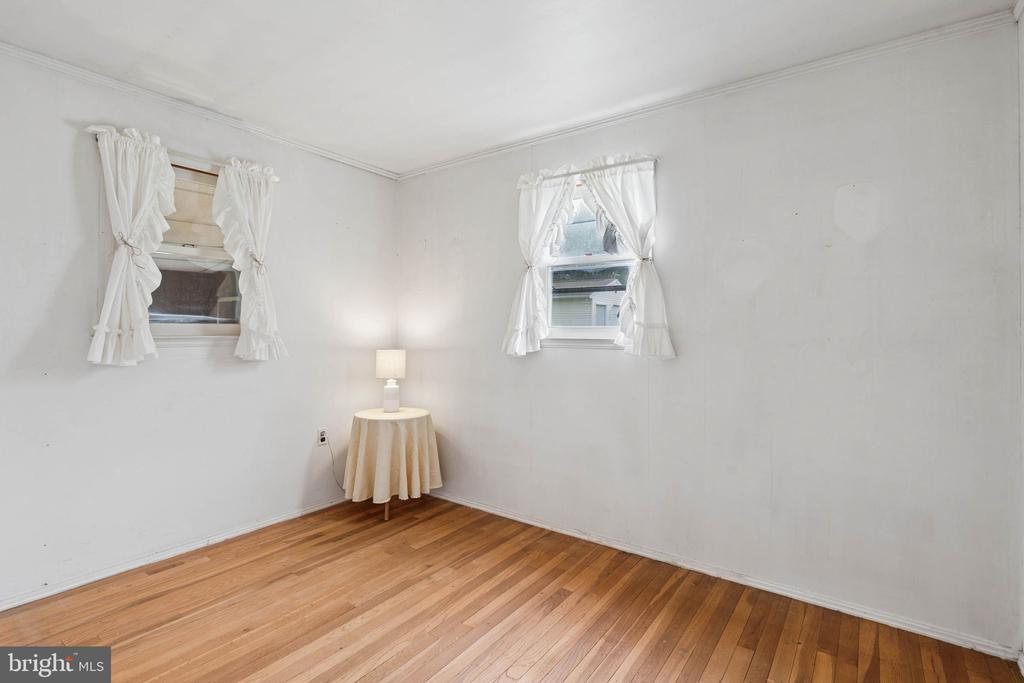 Sparse room with hardwood floors, two curtained windows, and a small draped table with a lamp.