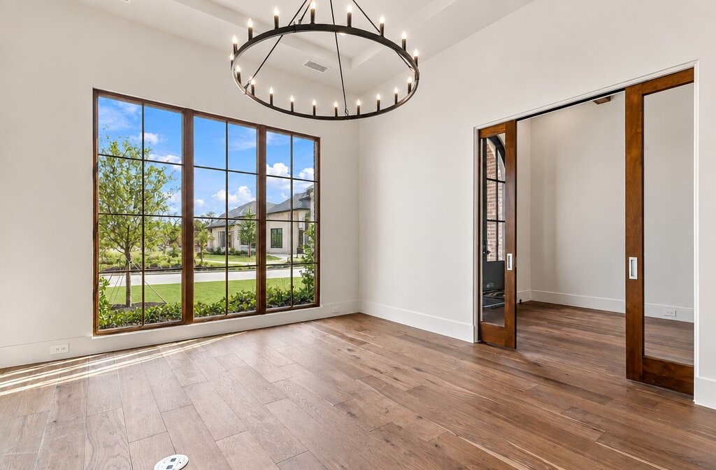 Empty dining room with a circular chandelier, wood floors, and a large window overlooking the street.