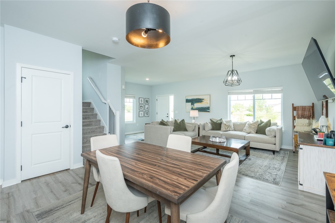 Open-plan dining and living area with a wood table, gray sectional, staircase, and large windows.
