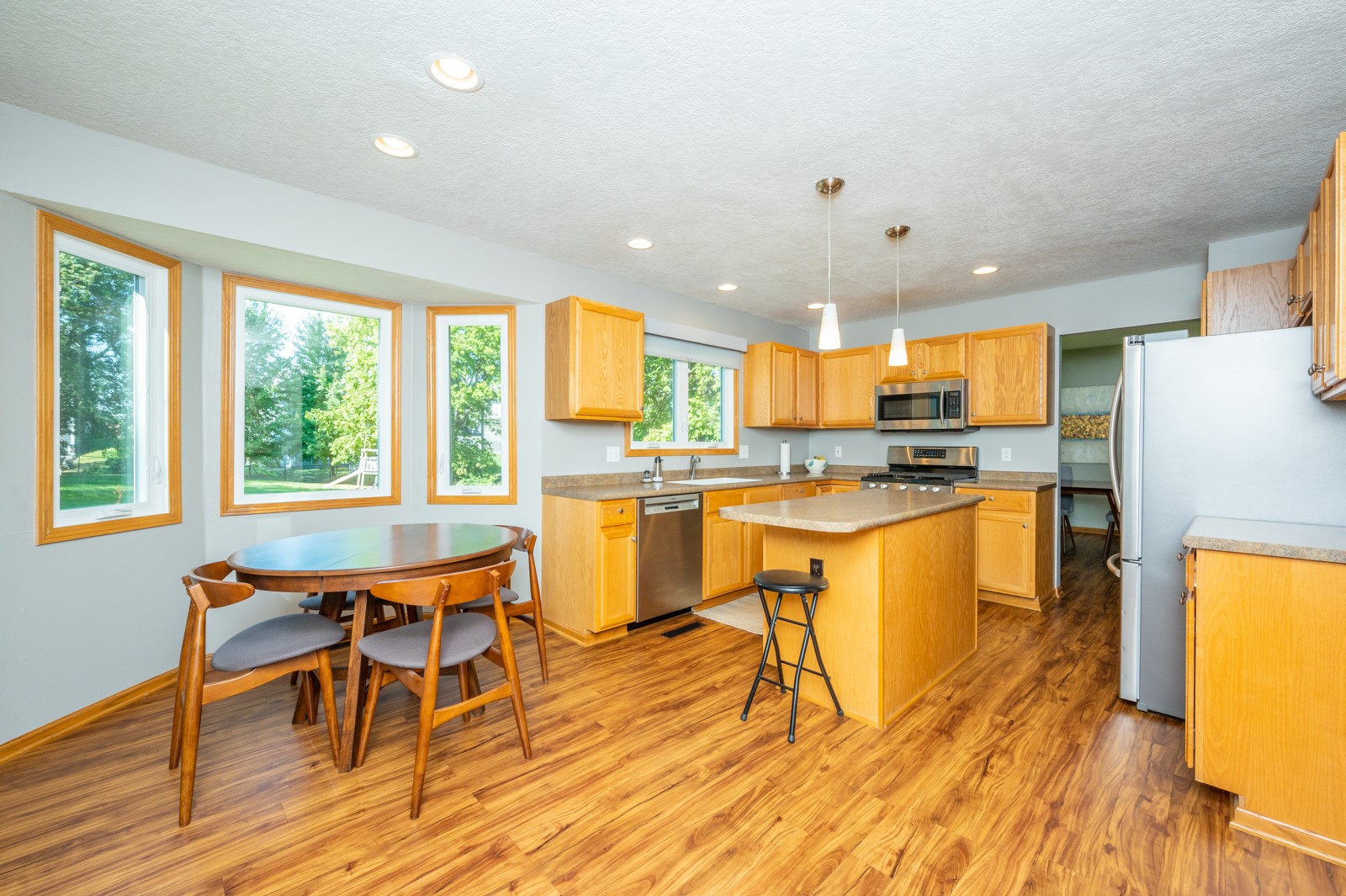 Kitchen with a central island, honey-colored cabinets, stainless steel appliances, and a round dining table by large windows.