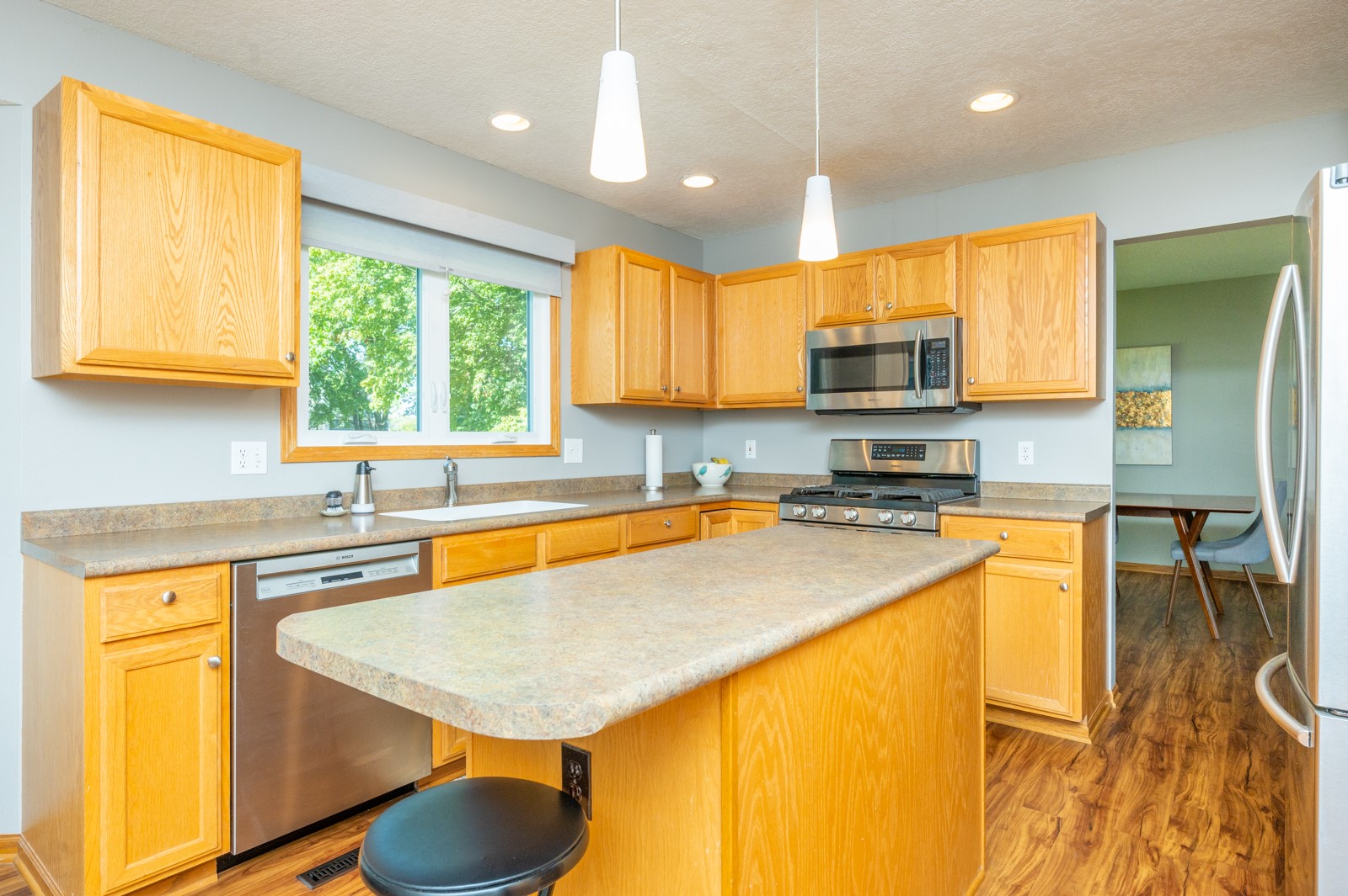 Kitchen with a large island, honey-colored cabinets, stainless steel appliances, and a window overlooking trees.