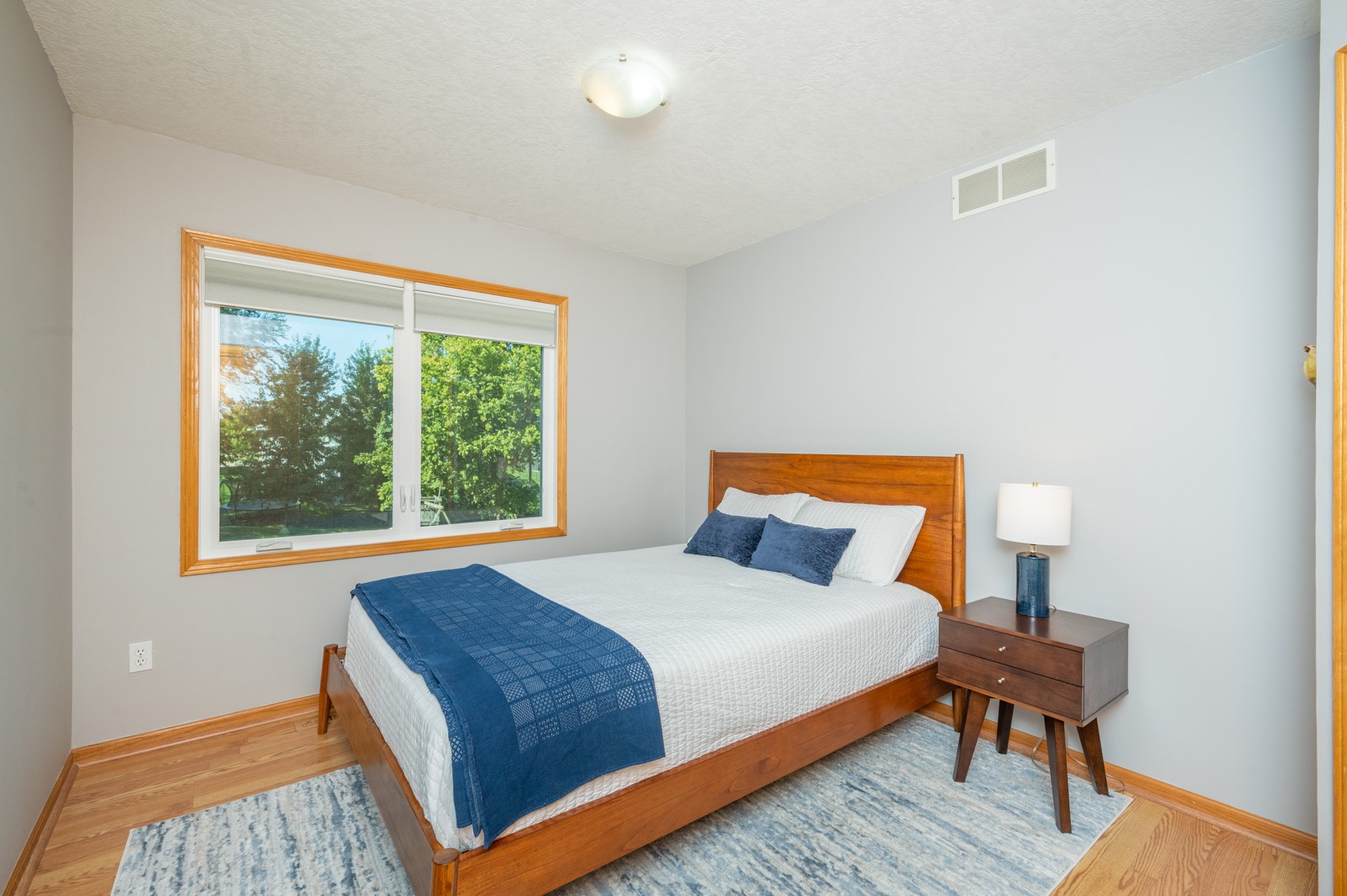 Bedroom with a wooden bed, blue accents, hardwood floors, and a large window overlooking trees.