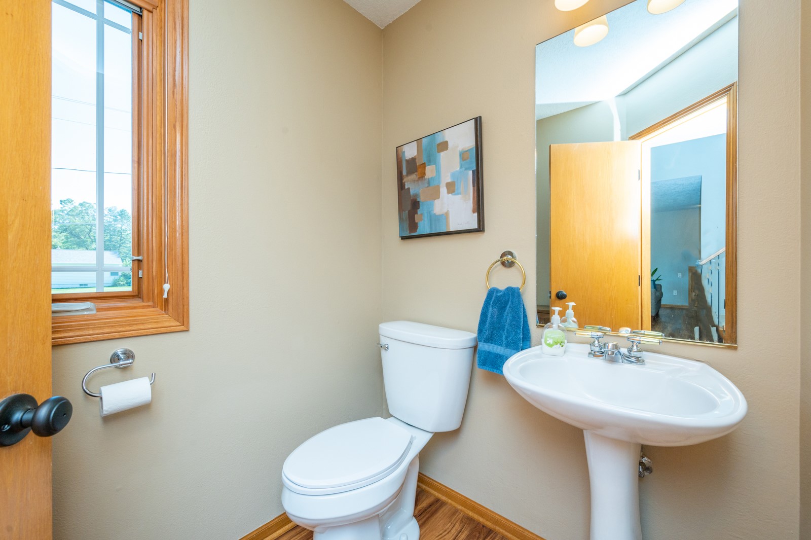Bathroom with a white pedestal sink, toilet, large mirror, and wood-trimmed window.