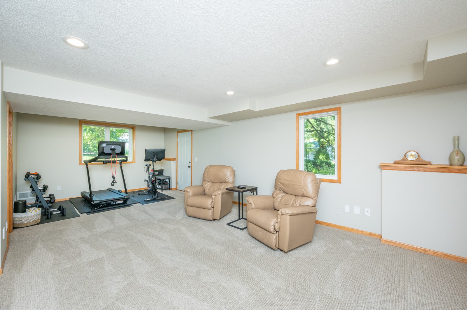 Basement exercise room with a treadmill, stationary bike, dumbbells, and two tan leather recliners.