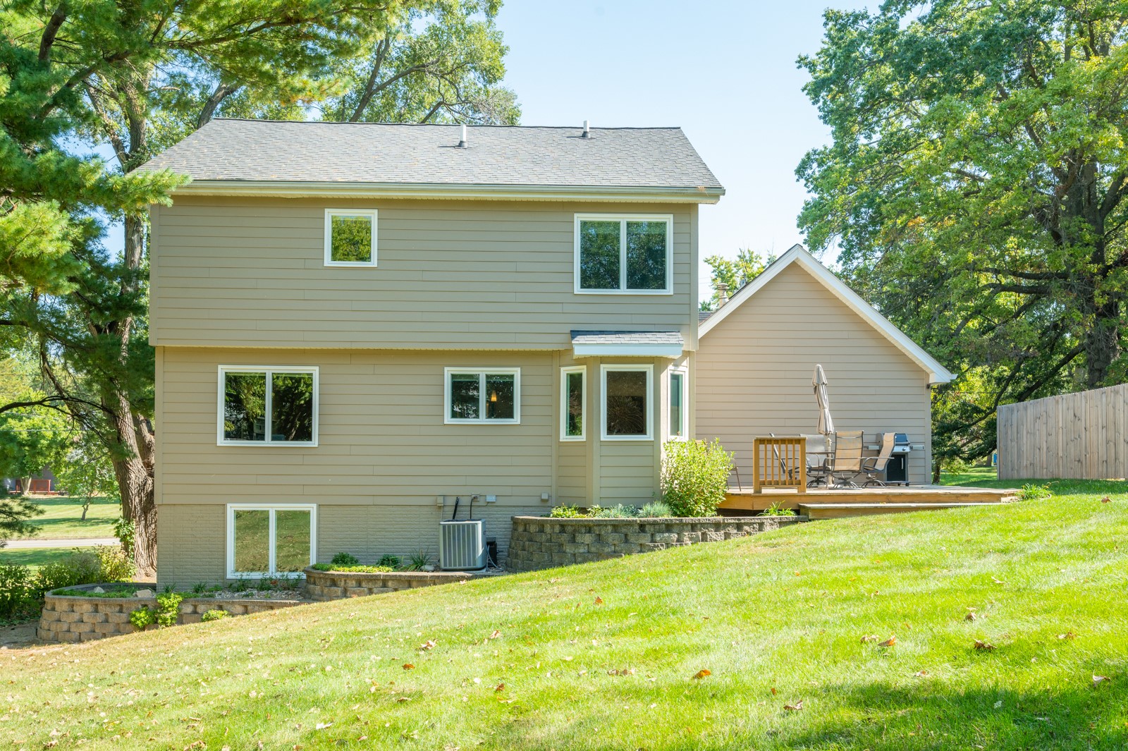 Two-story beige house with a raised deck, patio furniture, and a sloping lawn.