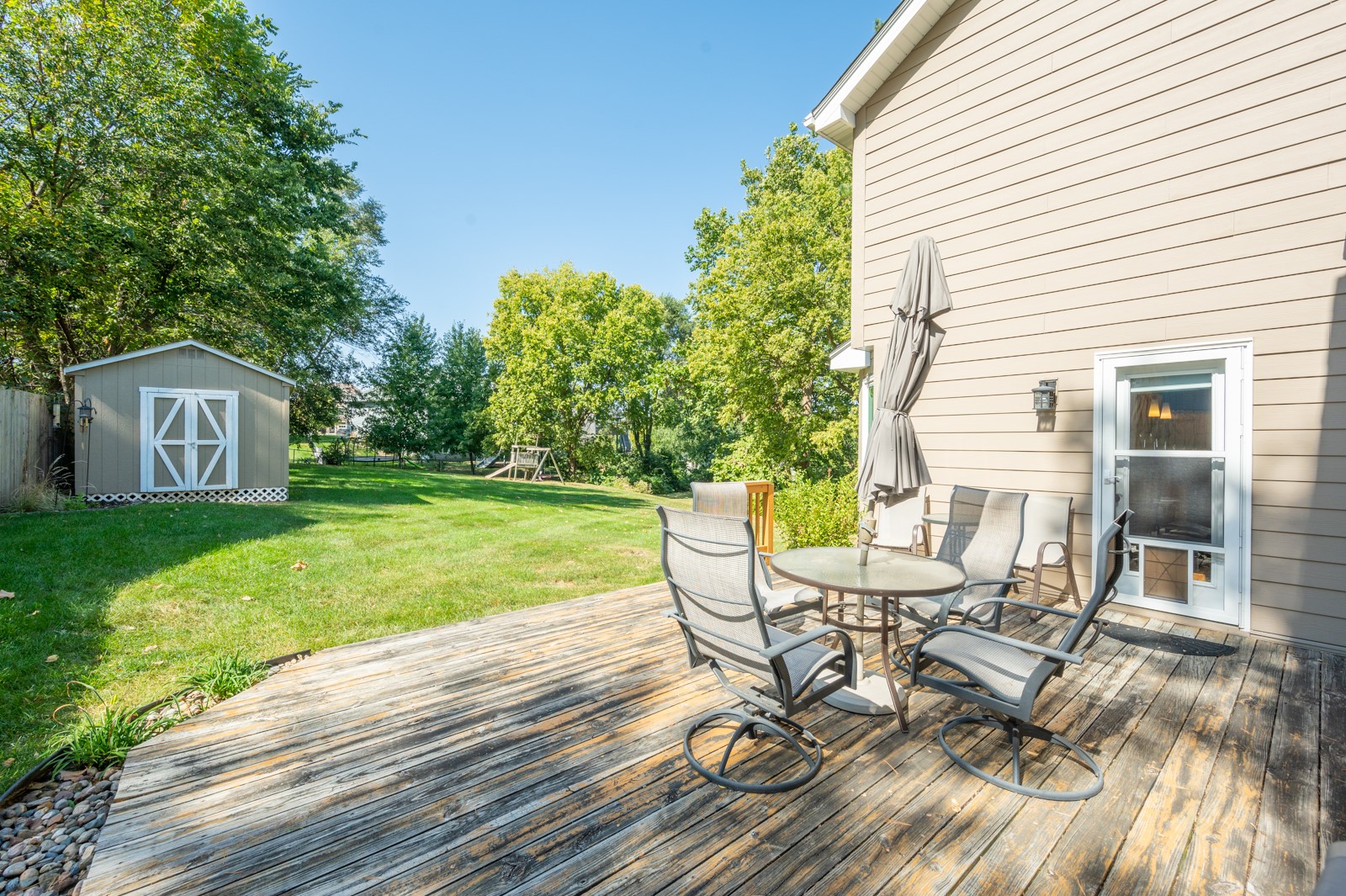 Backyard deck with patio dining set overlooking a lawn, trees, storage shed, and swing set.