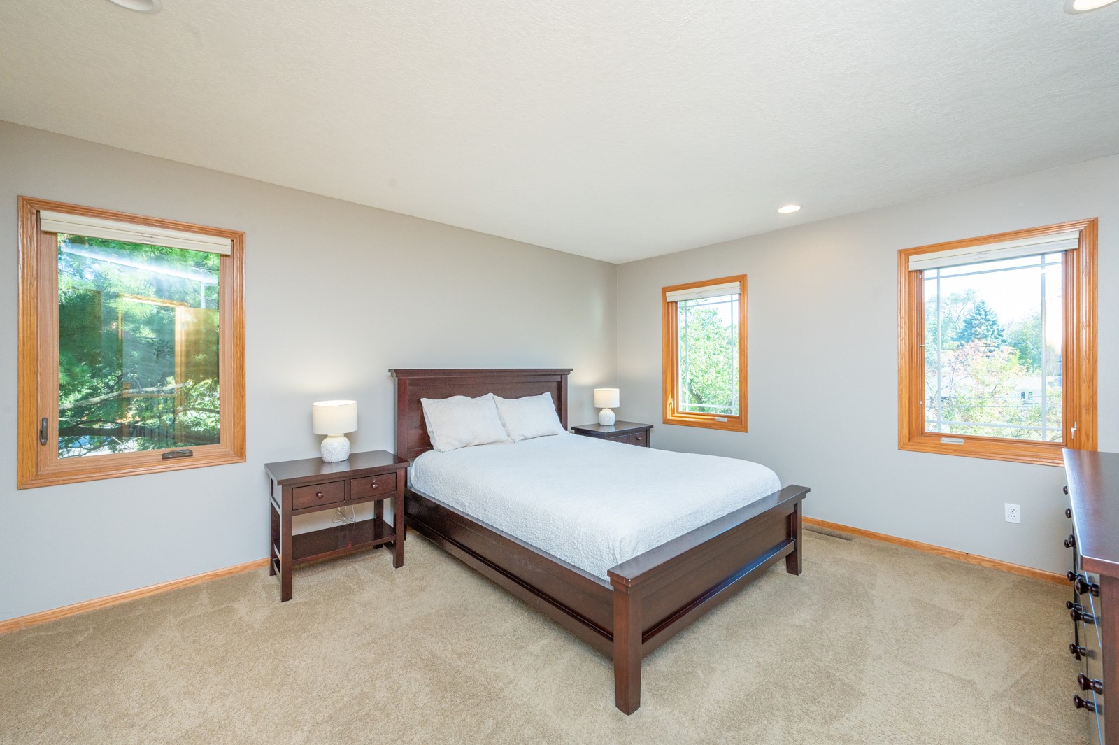 Bedroom with a dark wood bed, white bedding, beige carpet, and three windows.