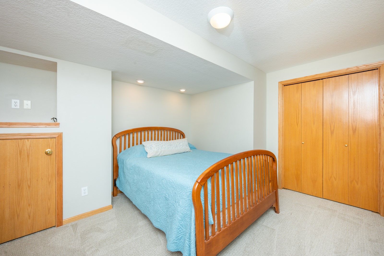 Bedroom with a wooden bed, light blue quilt, beige carpet, and wood-paneled closet doors.