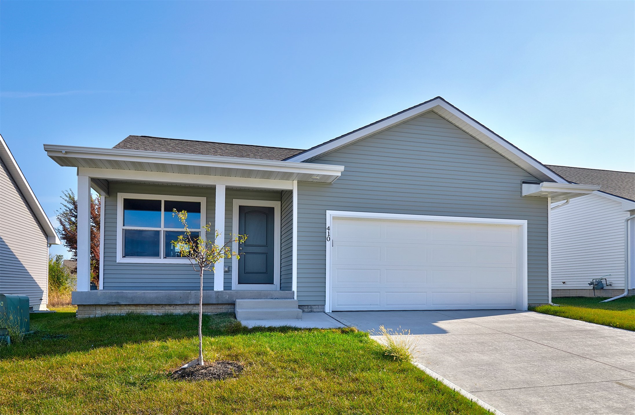 Gray-sided single-story home with a covered porch, young front-yard tree, and attached garage.