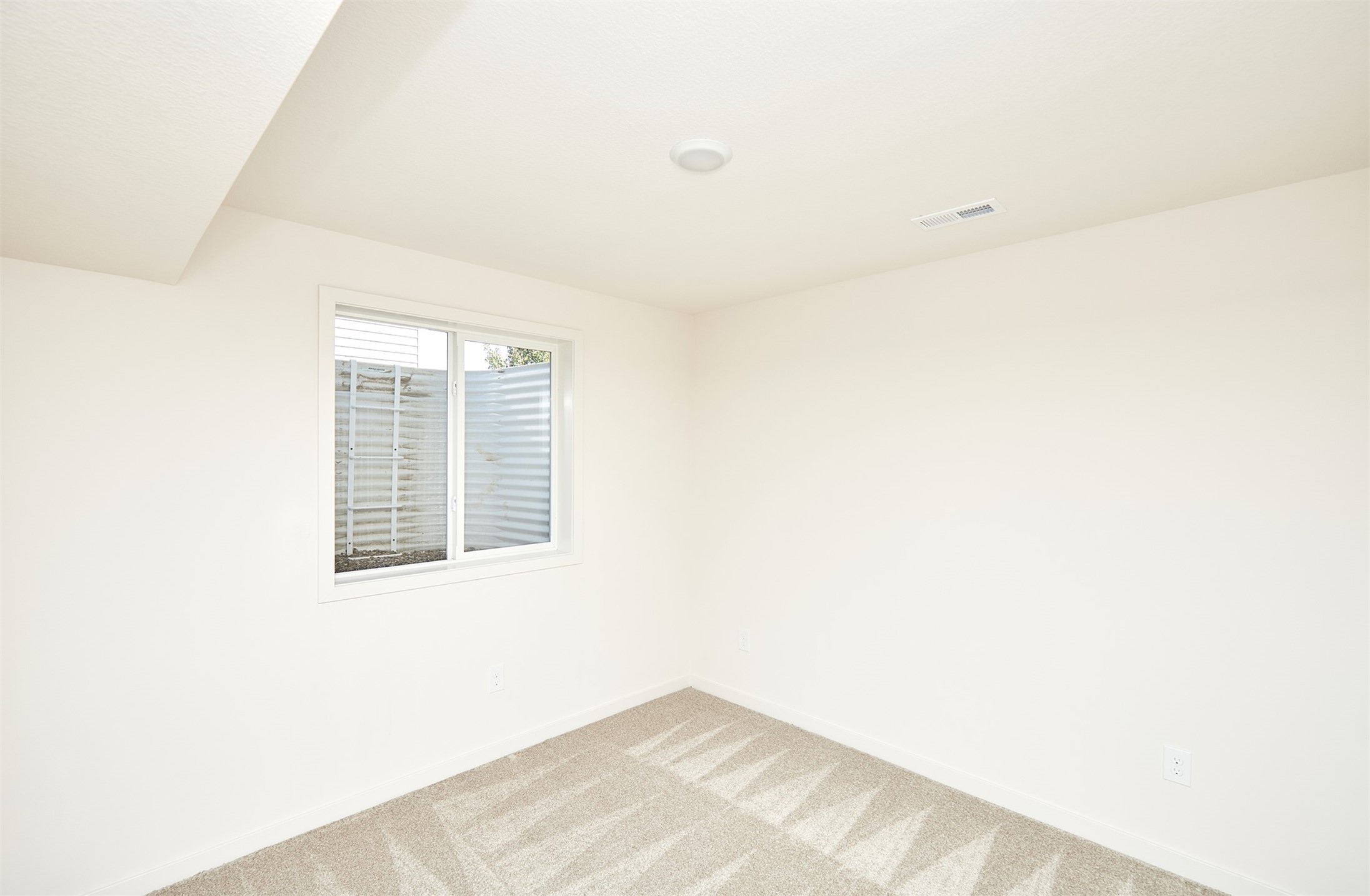 Empty carpeted room with white walls and a window overlooking a fence.