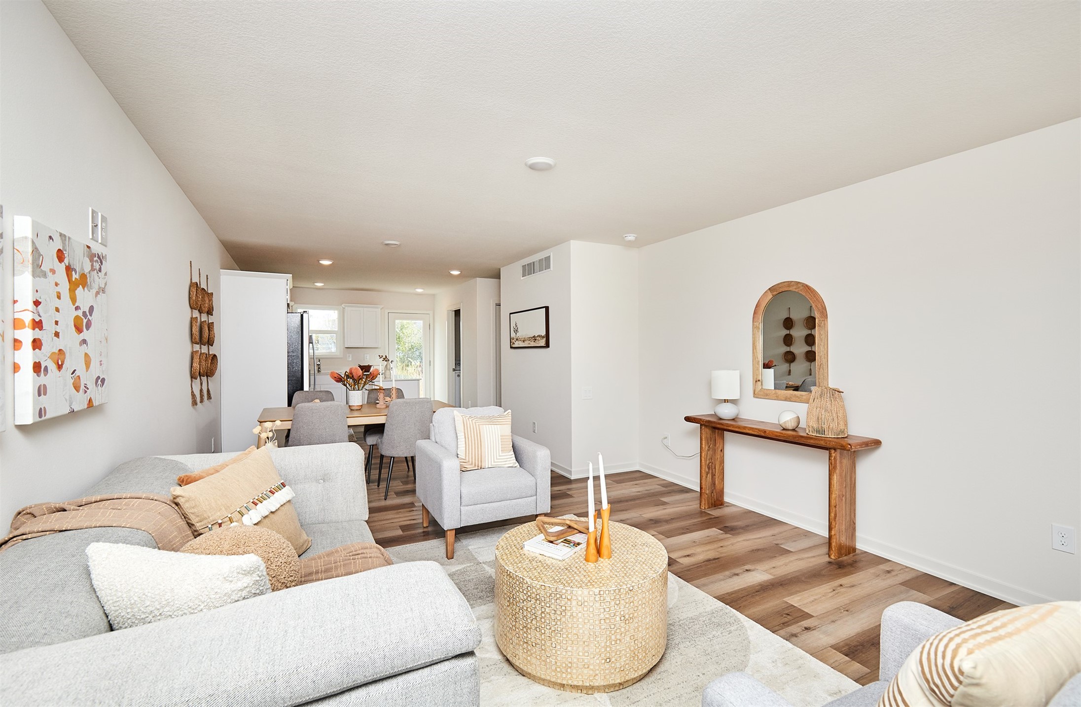 Open-plan living room with gray sofas, woven coffee table, wood flooring, and dining area leading to the kitchen.