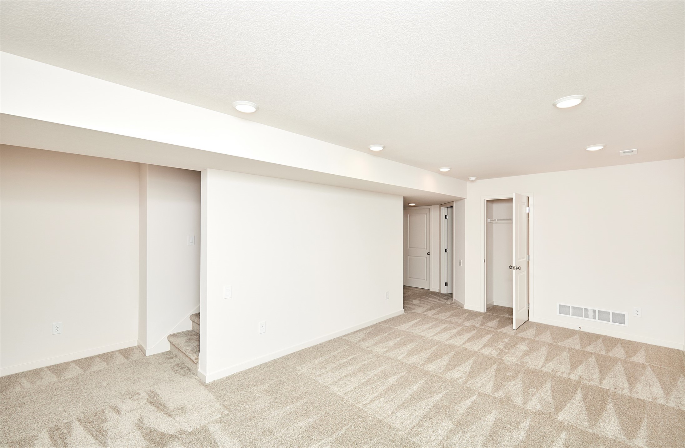 Finished basement room with beige patterned carpet, recessed lights, stairs, and an open closet.