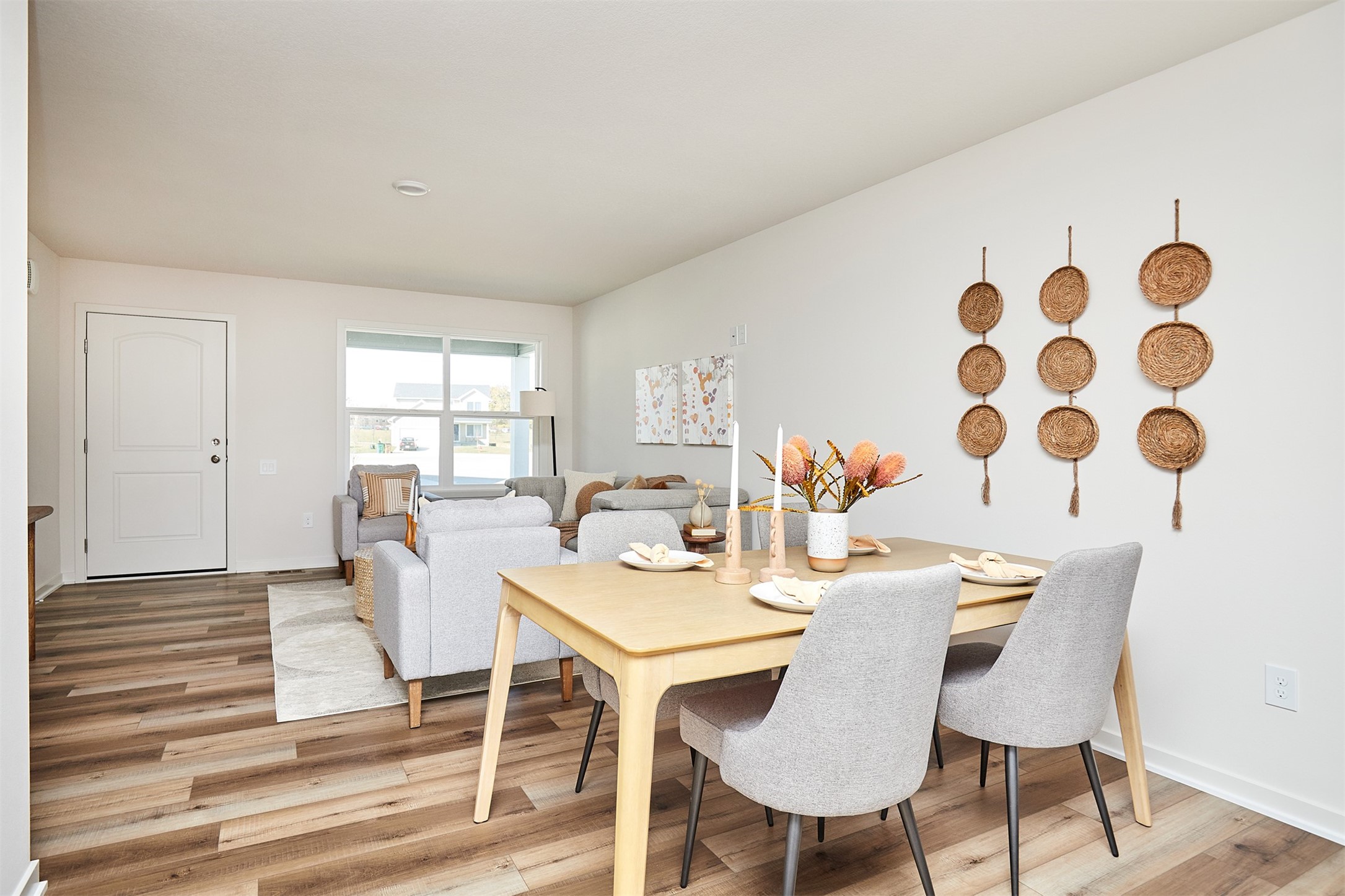 Open-plan dining and living area with a light wood table, gray seating, and woven wall decor.