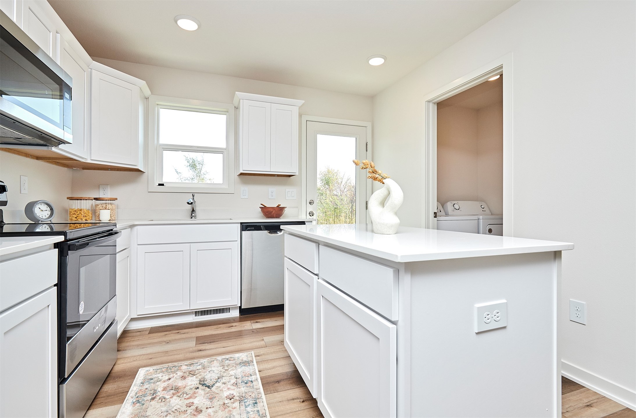 Bright white kitchen with a large island, stainless steel appliances, and a laundry area through the doorway.