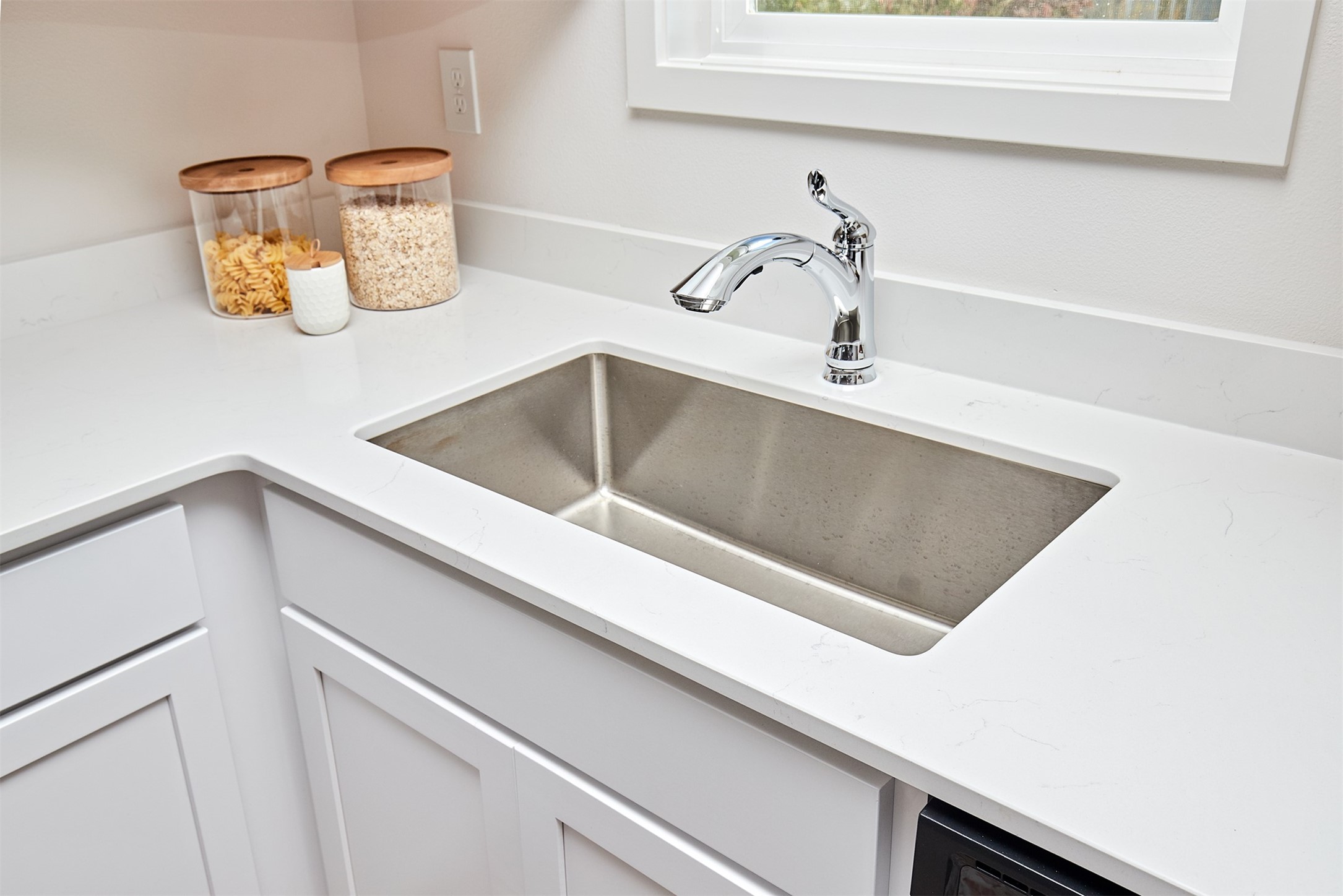 Kitchen sink with a chrome faucet, white counters, and jars of pasta and oats.