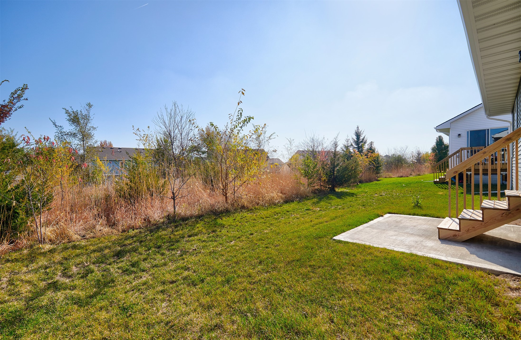Backyard with a wide green lawn, autumn shrubs, and wooden deck stairs beside a concrete patio.