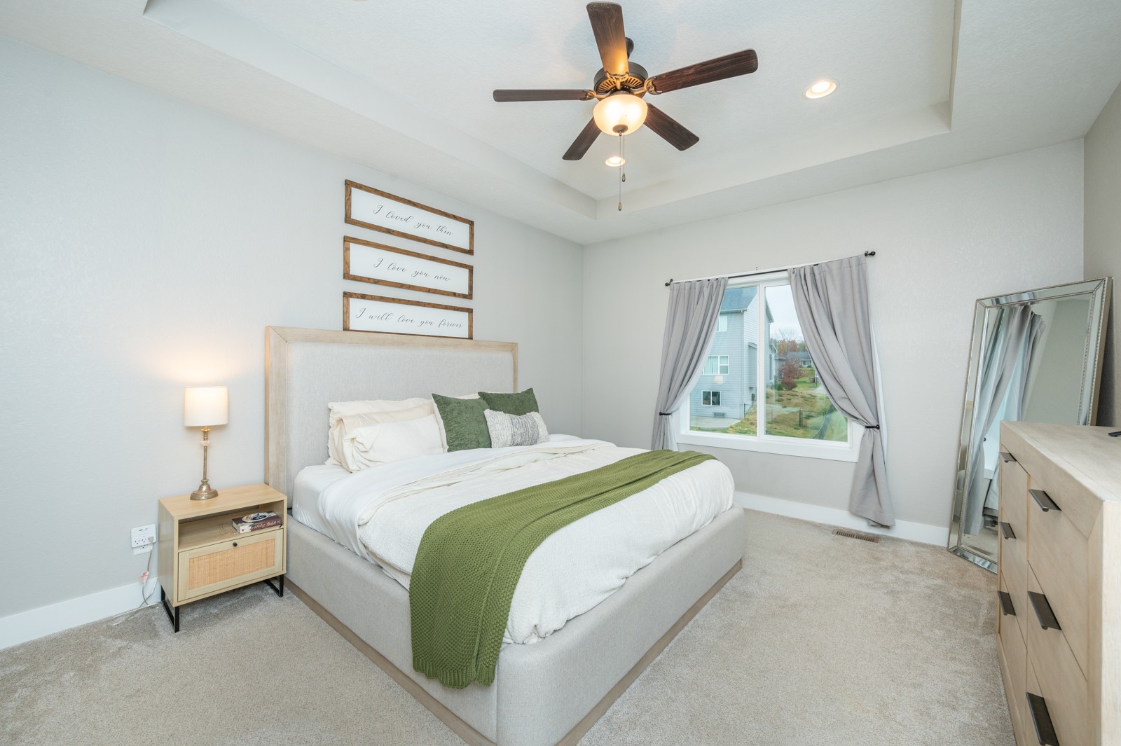 Staged bedroom with a light upholstered bed, green throw, curtained window, and ceiling fan.
