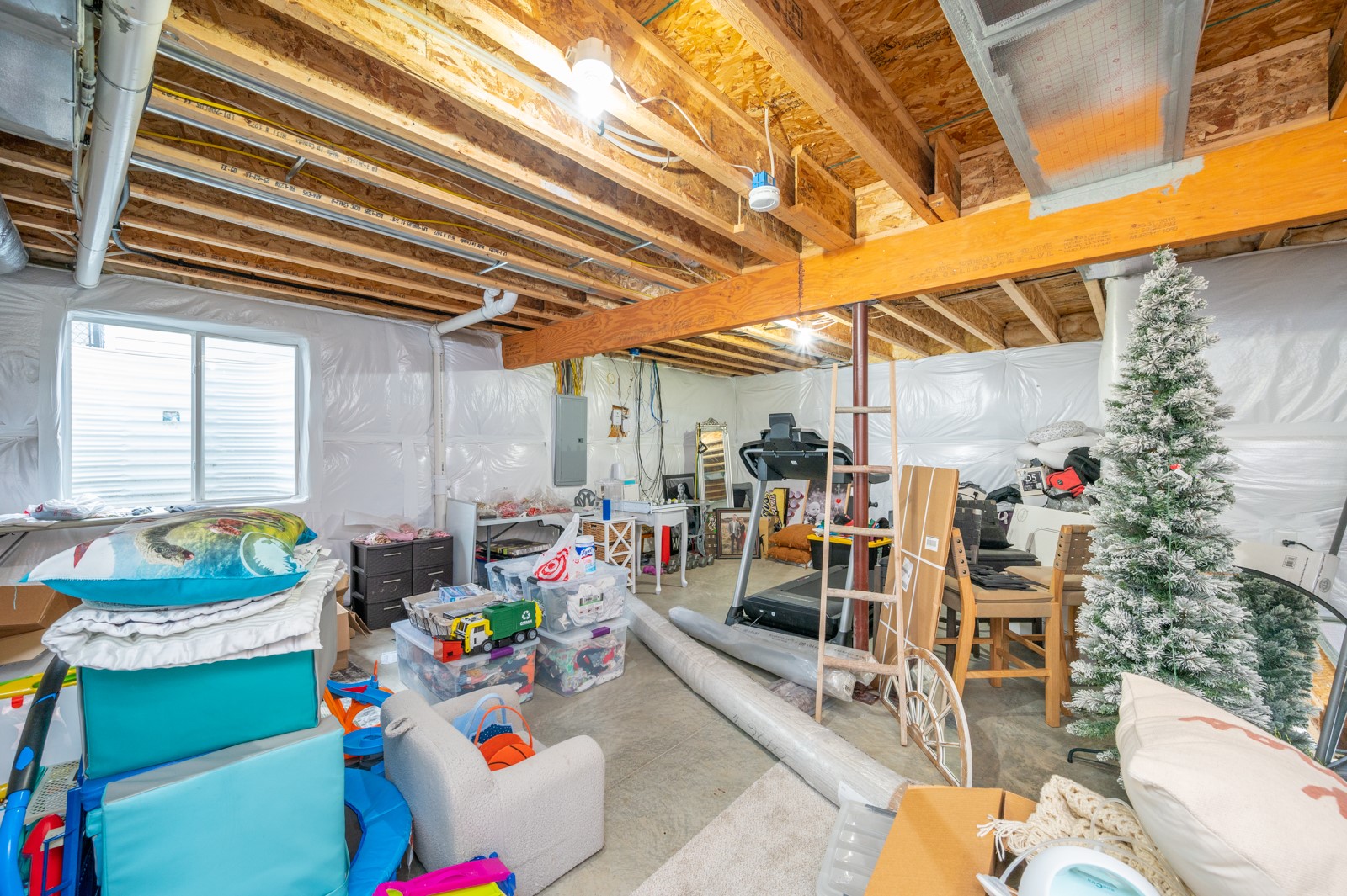 Unfinished basement with exposed ceiling joists, stored belongings, exercise equipment, and a small window.