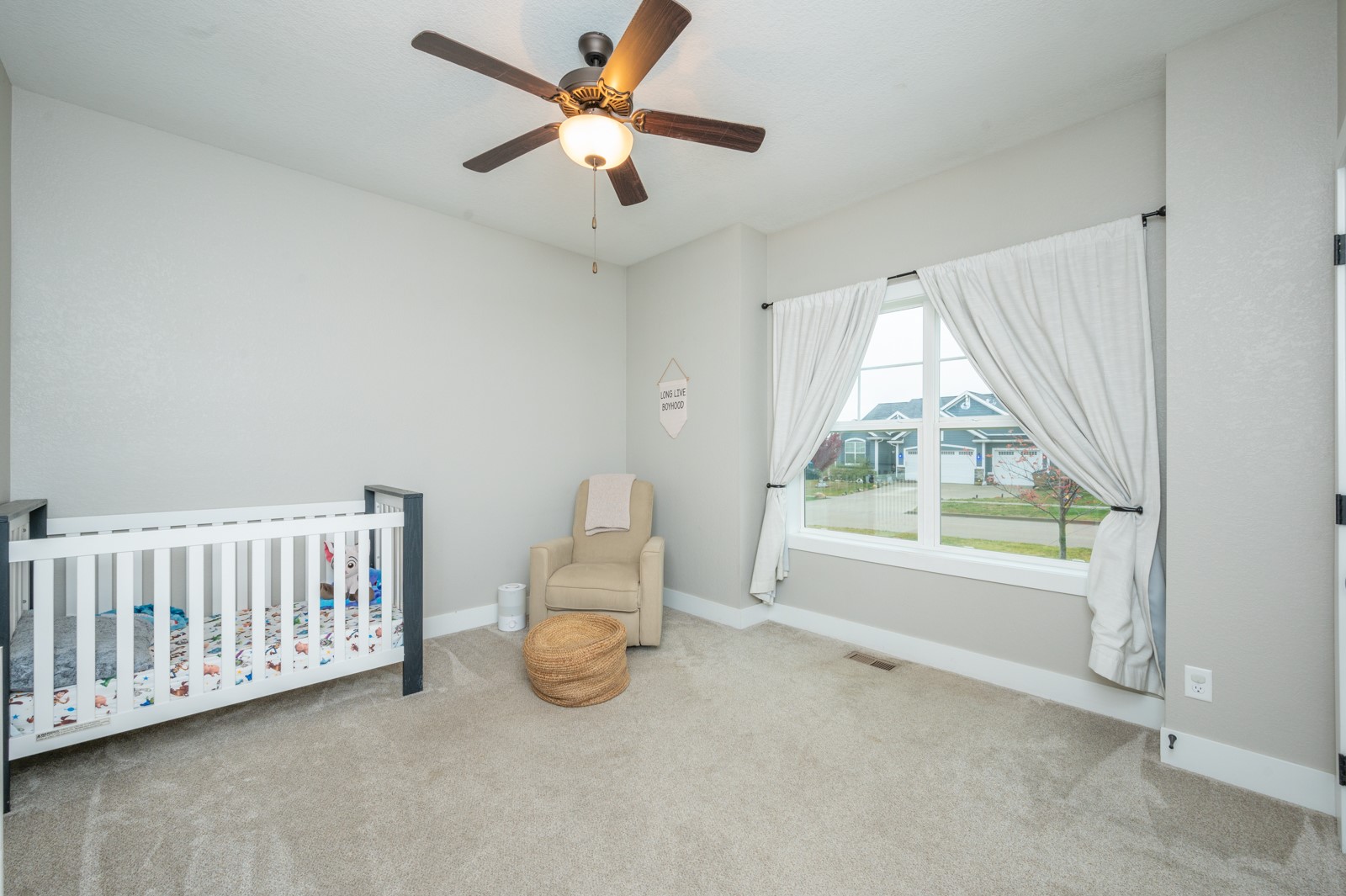 Nursery with a white crib, beige armchair, and large curtained window overlooking neighboring homes.