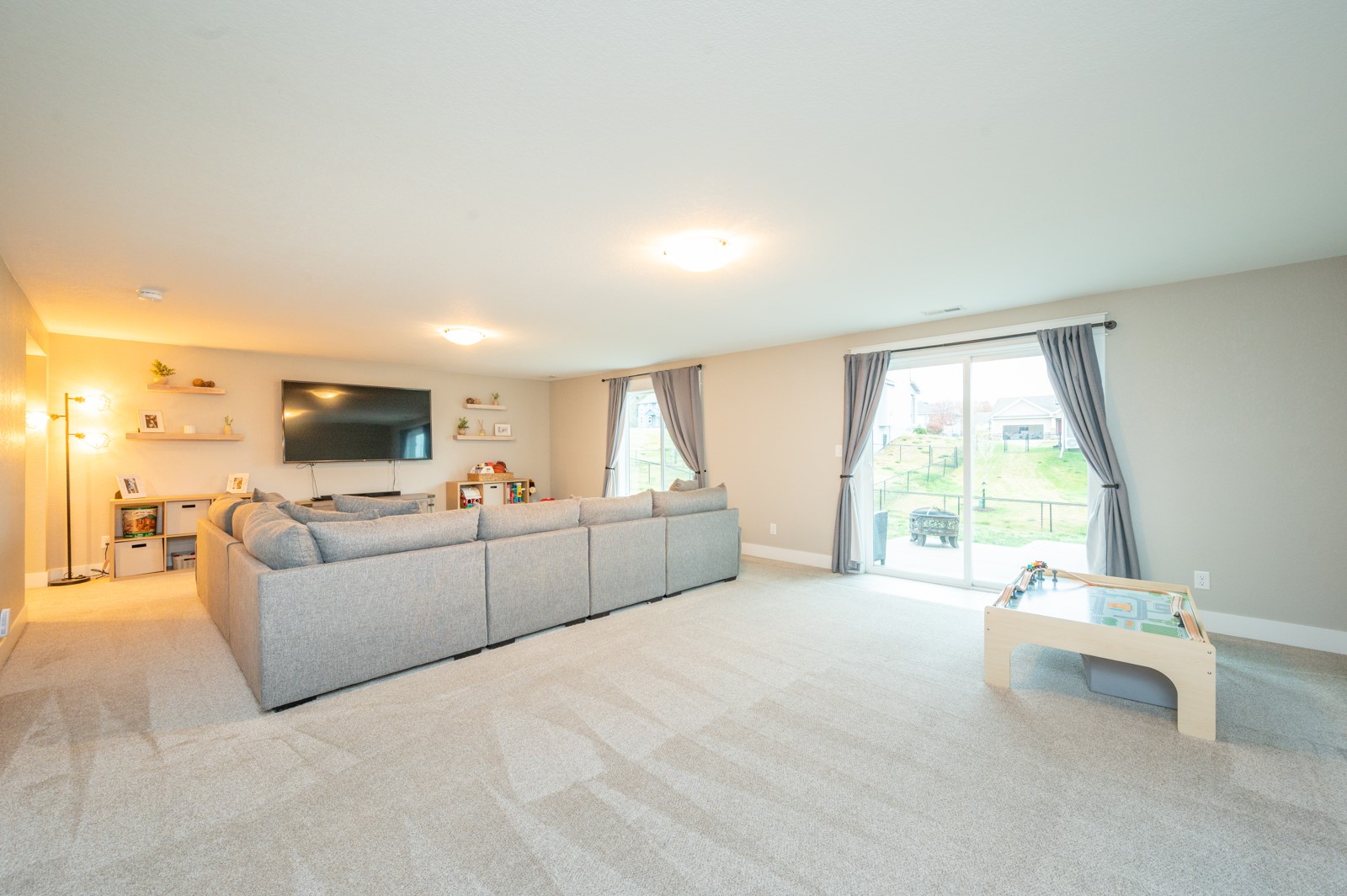 Furnished living room with a gray sectional, wall-mounted TV, sliding glass doors, and a small game table.