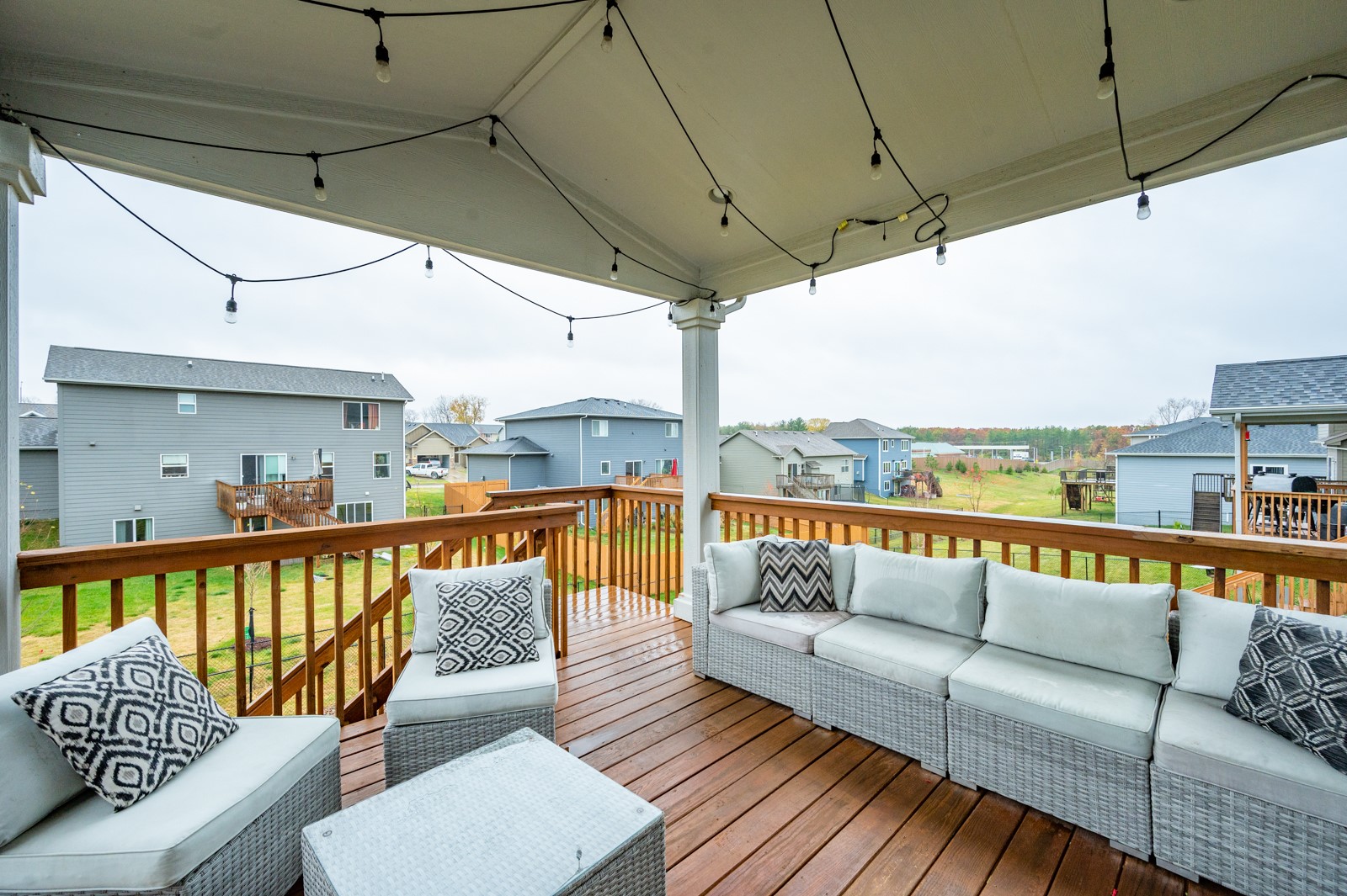 Covered wooden deck with white wicker seating and string lights overlooking neighboring homes.