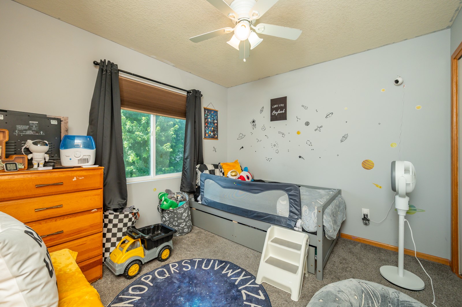 Children’s bedroom with a space-themed rug and wall decals, a gray bed, toys, and a window facing trees.