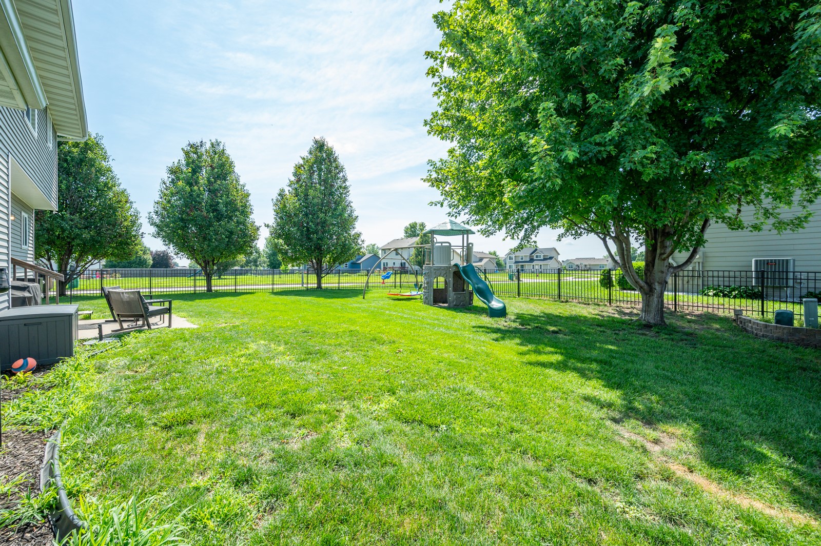 Fenced backyard with a play structure and slide, mature trees, and a wide green lawn.