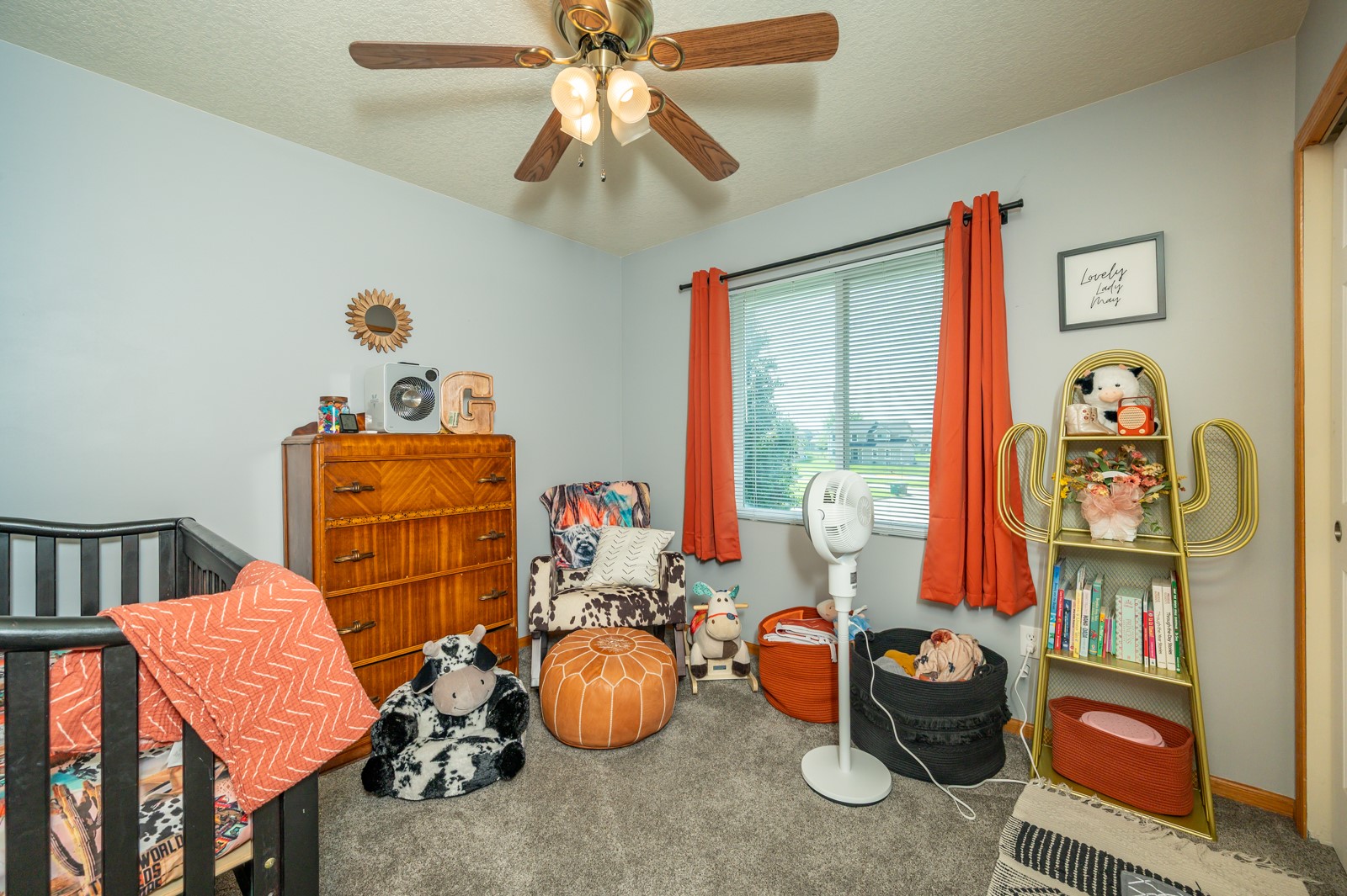 Furnished nursery with a black crib, coral curtains, wooden dresser, patterned chair, and children’s toys.