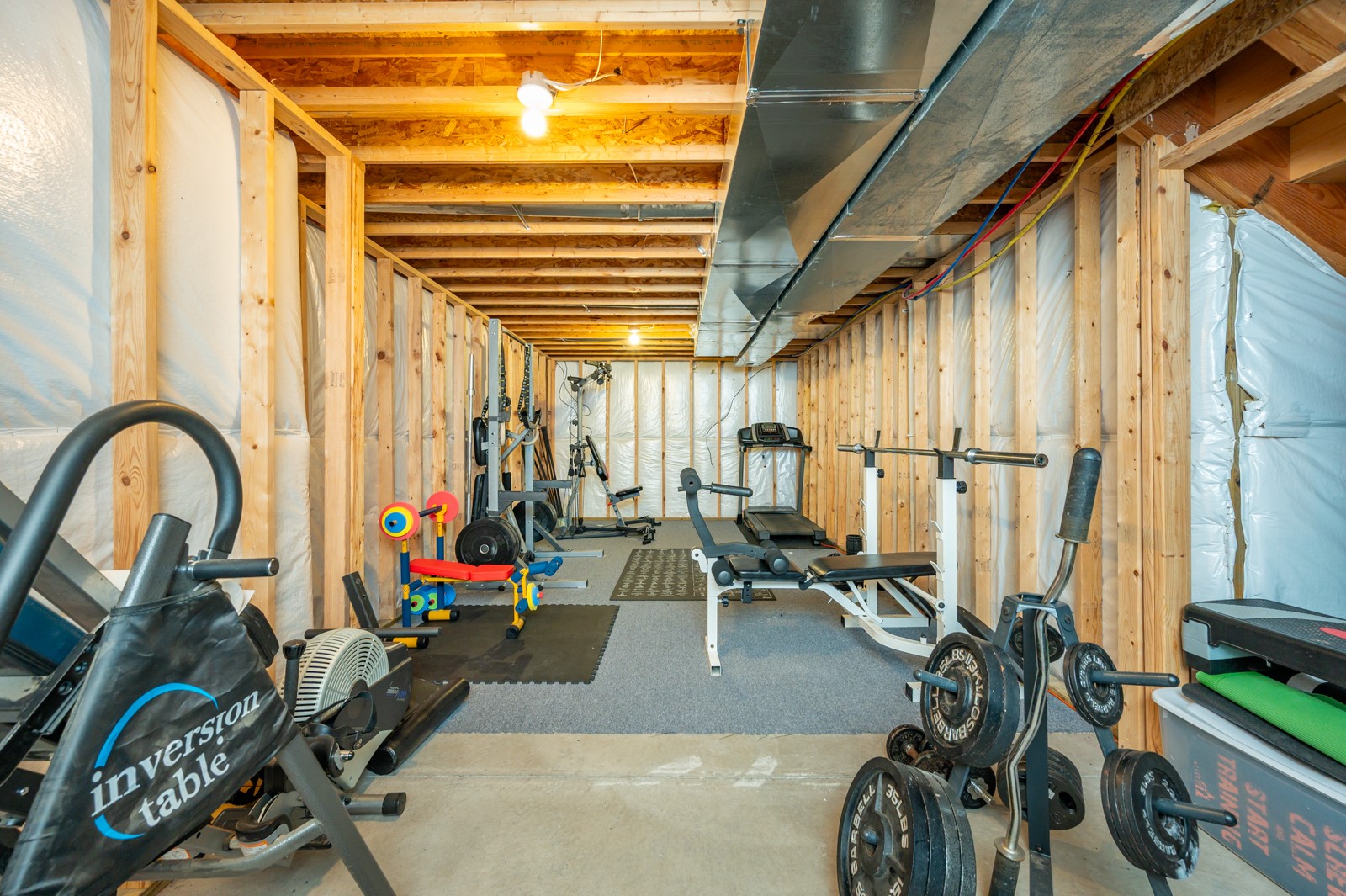 Basement home gym with exercise equipment, exposed framing and overhead ductwork.