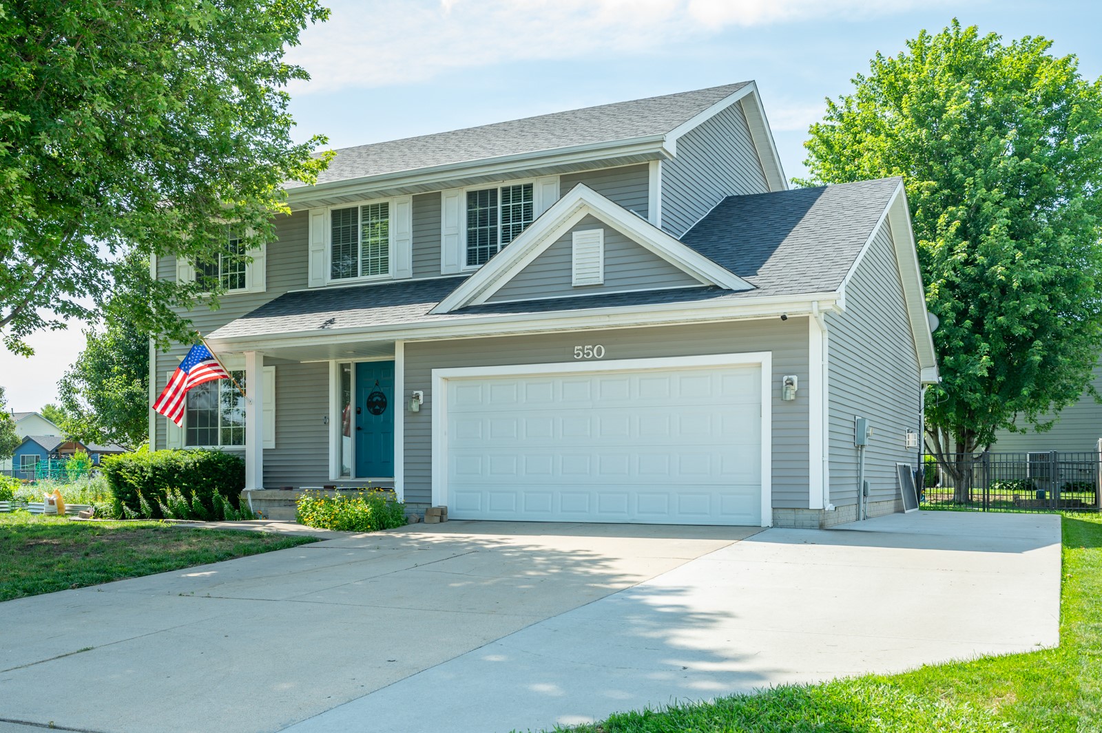 Two-story gray house with a teal entry door, double garage, U.S. flag, and wide concrete driveway.