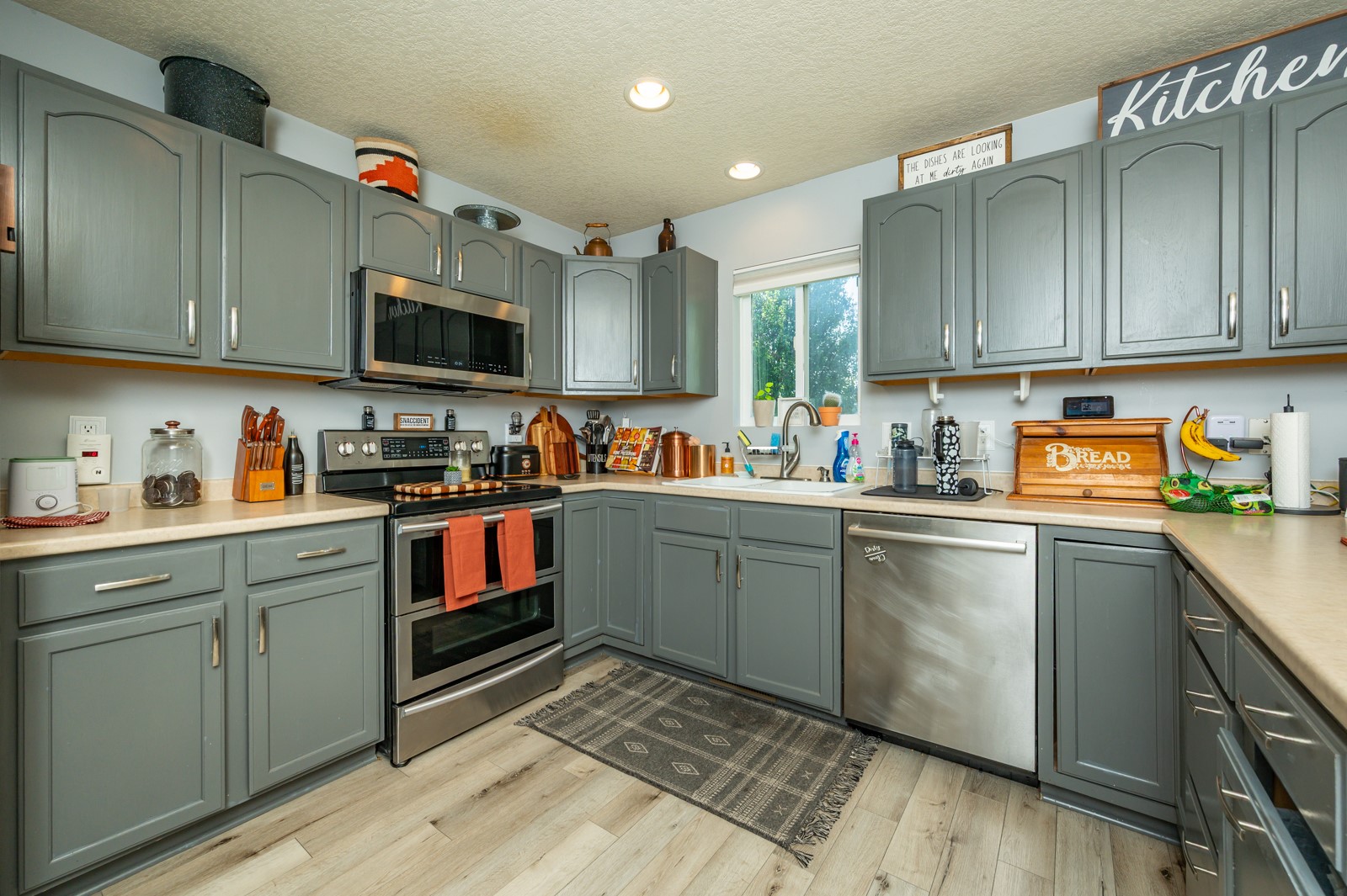Kitchen with gray-green cabinets, stainless steel appliances, and a sink beneath a window.