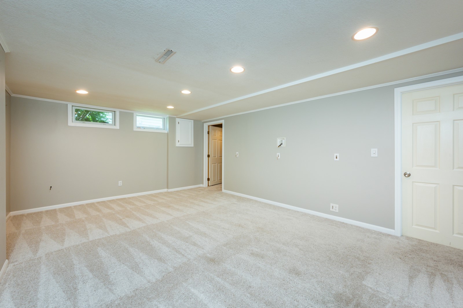 Empty carpeted basement room with recessed lights and two high windows.